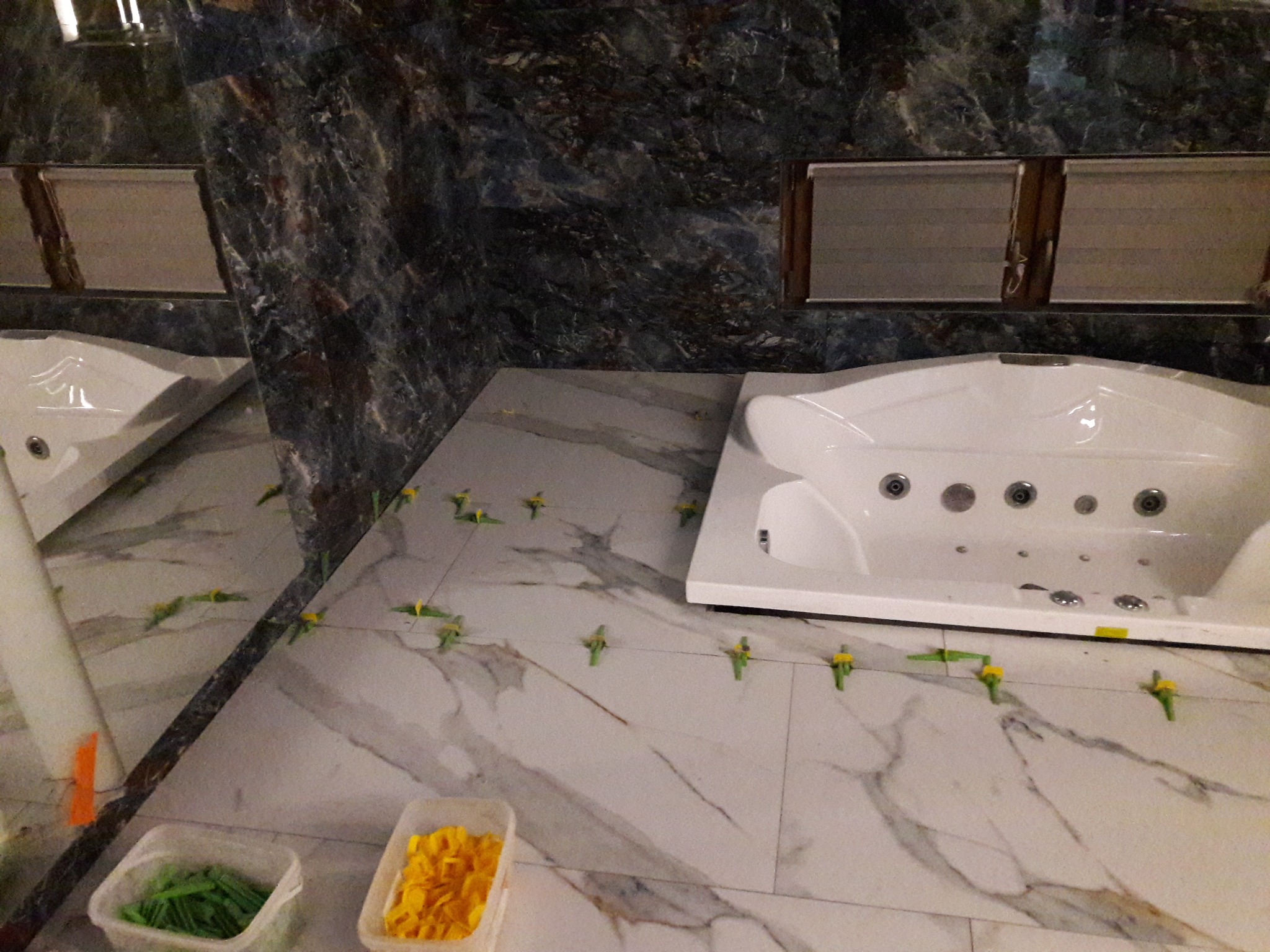 Układanie marmurowych płytek podłogowych w łazience z wanną z hydromasażem, widoczne plastikowe kliny i pojemniki z elementami dystansowymi.