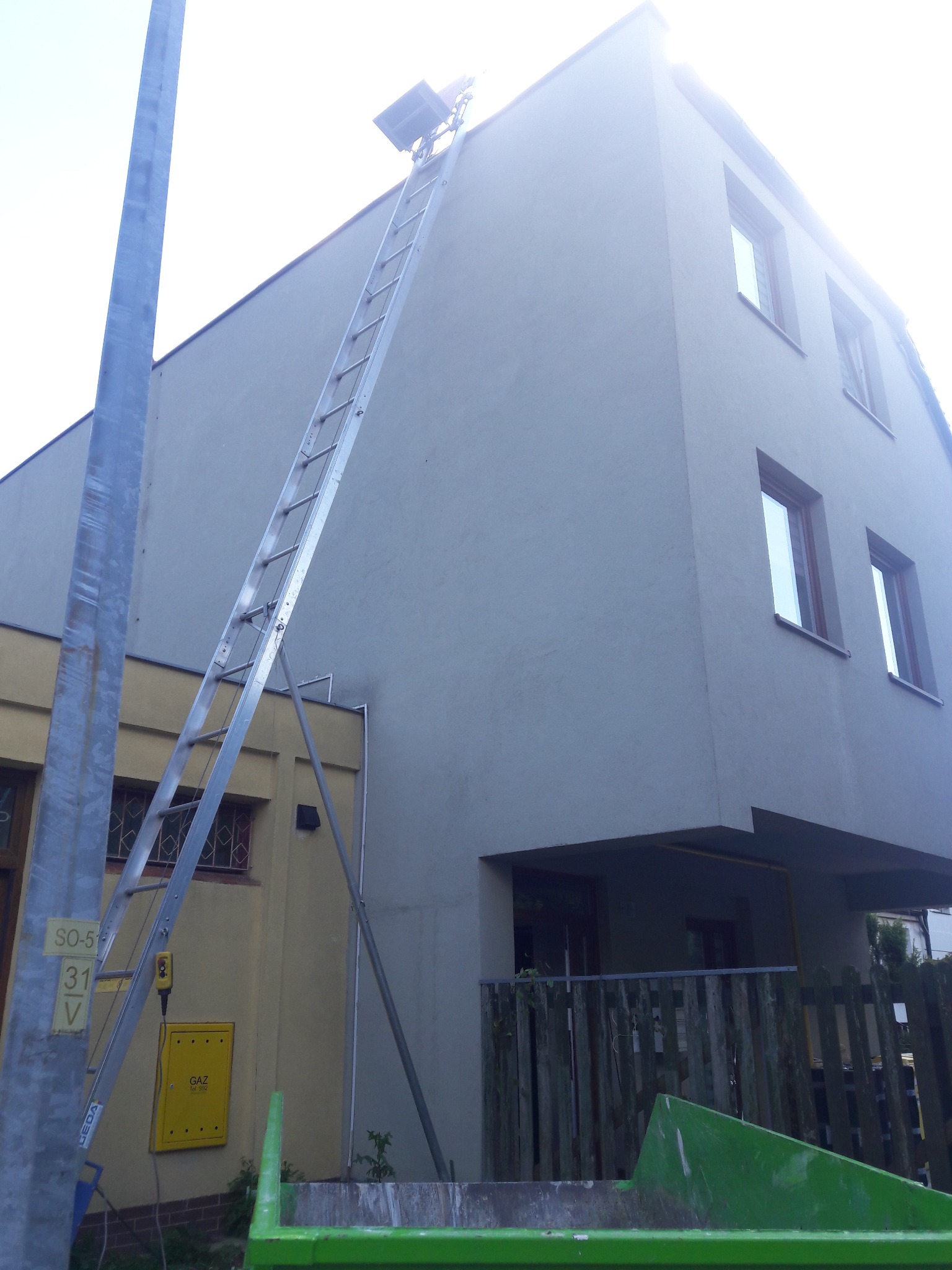 Podnośnik dekarski ustawiony przy szarej ścianie budynku, widoczny kontener na gruz i skrzynka gazowa na żółtej ścianie sąsiedniego budynku.