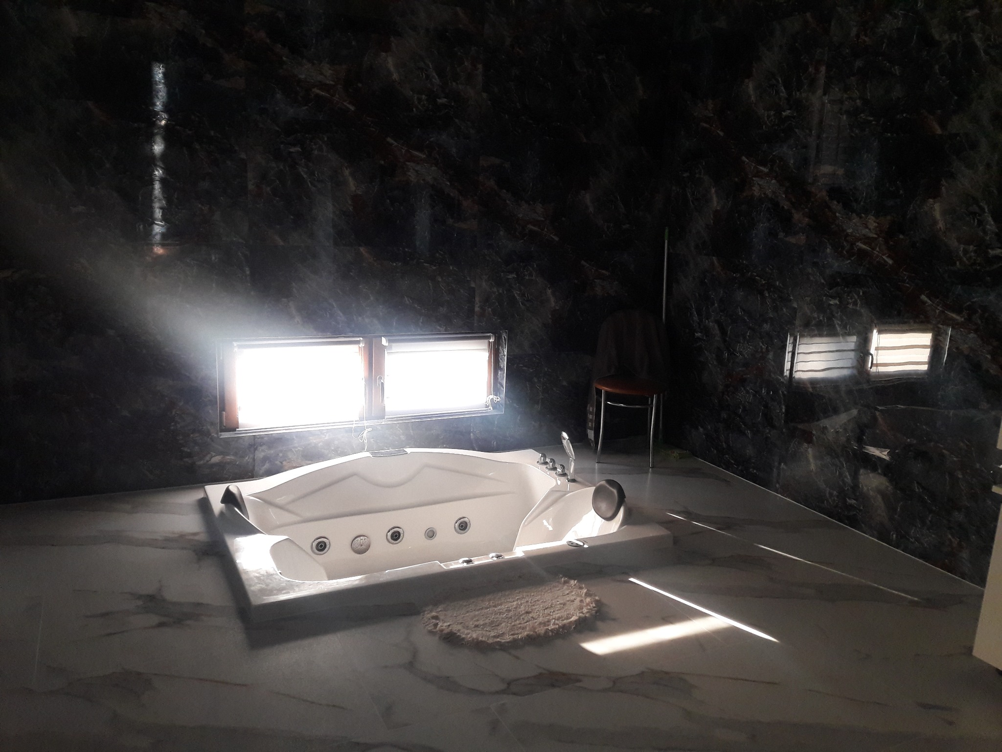 Biała wanna z hydromasażem na tle ciemnych marmurowych ścian, oświetlona promieniami słońca wpadającymi przez okno.