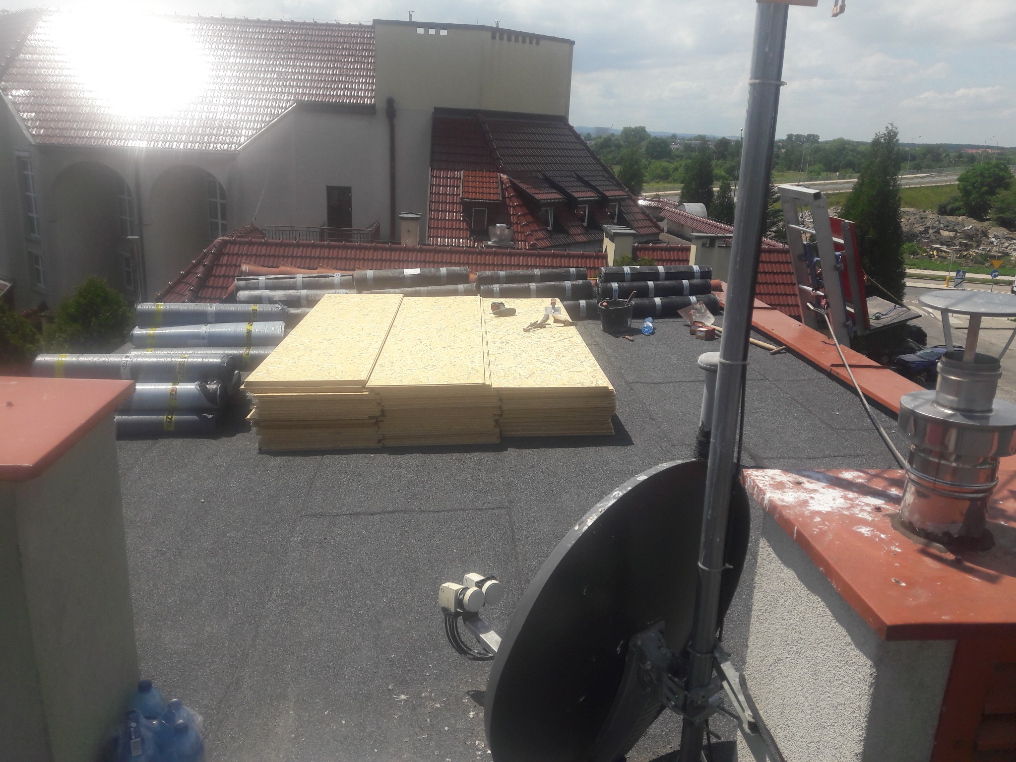 Przygotowanie do krycia dachu: rolki papy, płyty OSB i narzędzia na płaskim dachu budynku z widocznymi elementami wentylacji i anteną satelitarną.