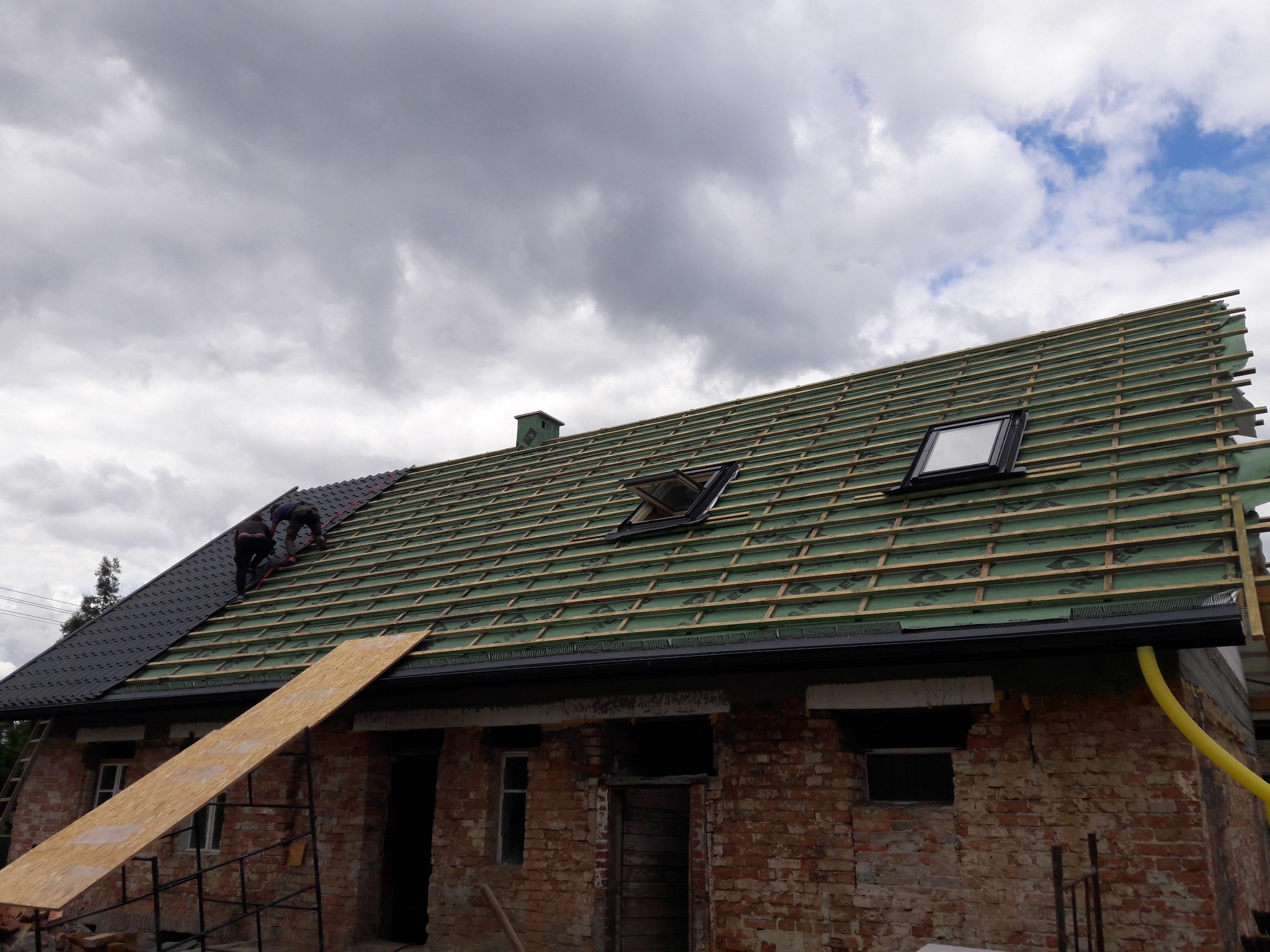 Częściowo pokryty dach z dwoma oknami dachowymi na tle zachmurzonego nieba, widoczni dekarze pracujący na dachu ceglanego budynku.