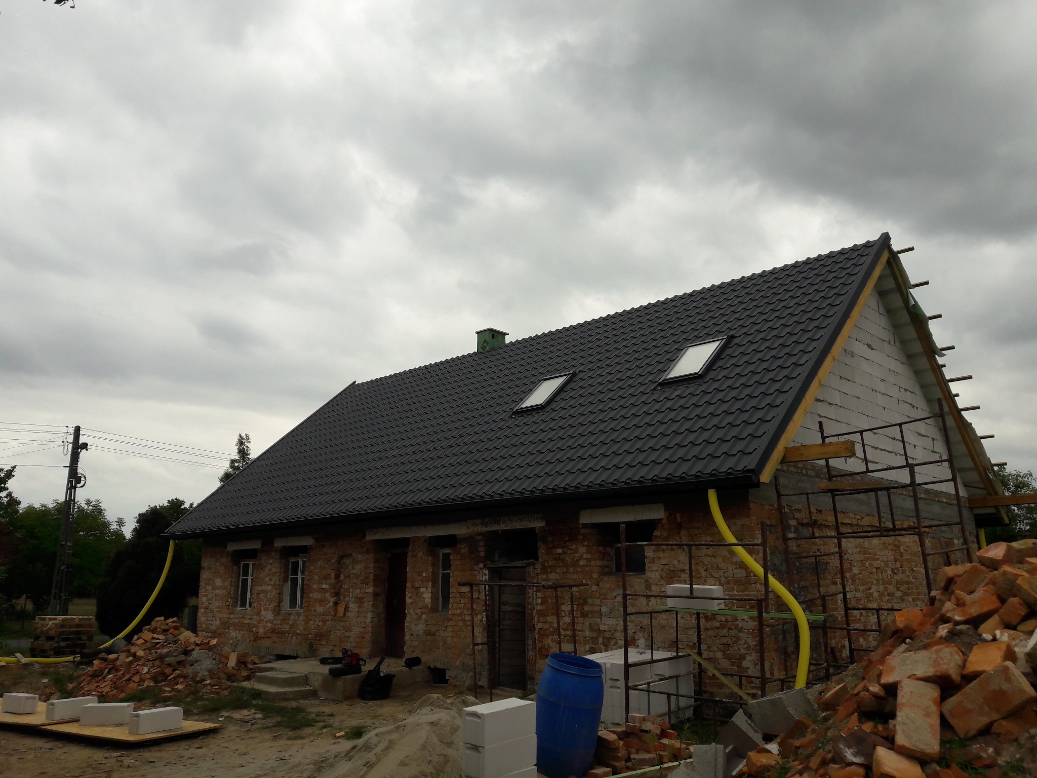 Budynek w trakcie remontu z częściowo położonym czarnym dachem z oknami dachowymi, widoczne rusztowania i materiały budowlane, cegły i pustaki, pochmurne niebo w tle.