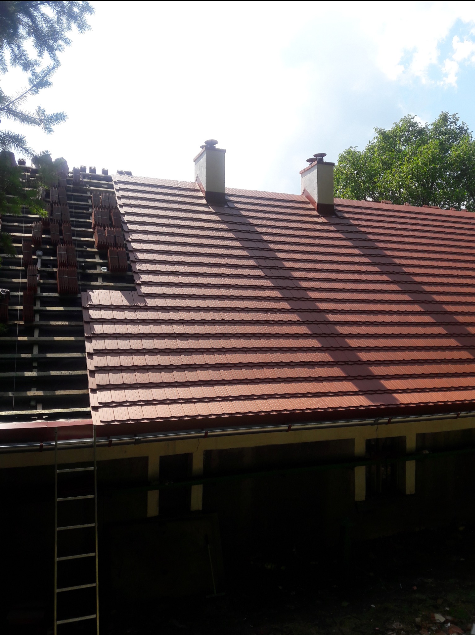 Częściowo pokryty dach z brązowej dachówki ceramicznej, widoczna więźba dachowa i kominy, drabina oparta o budynek