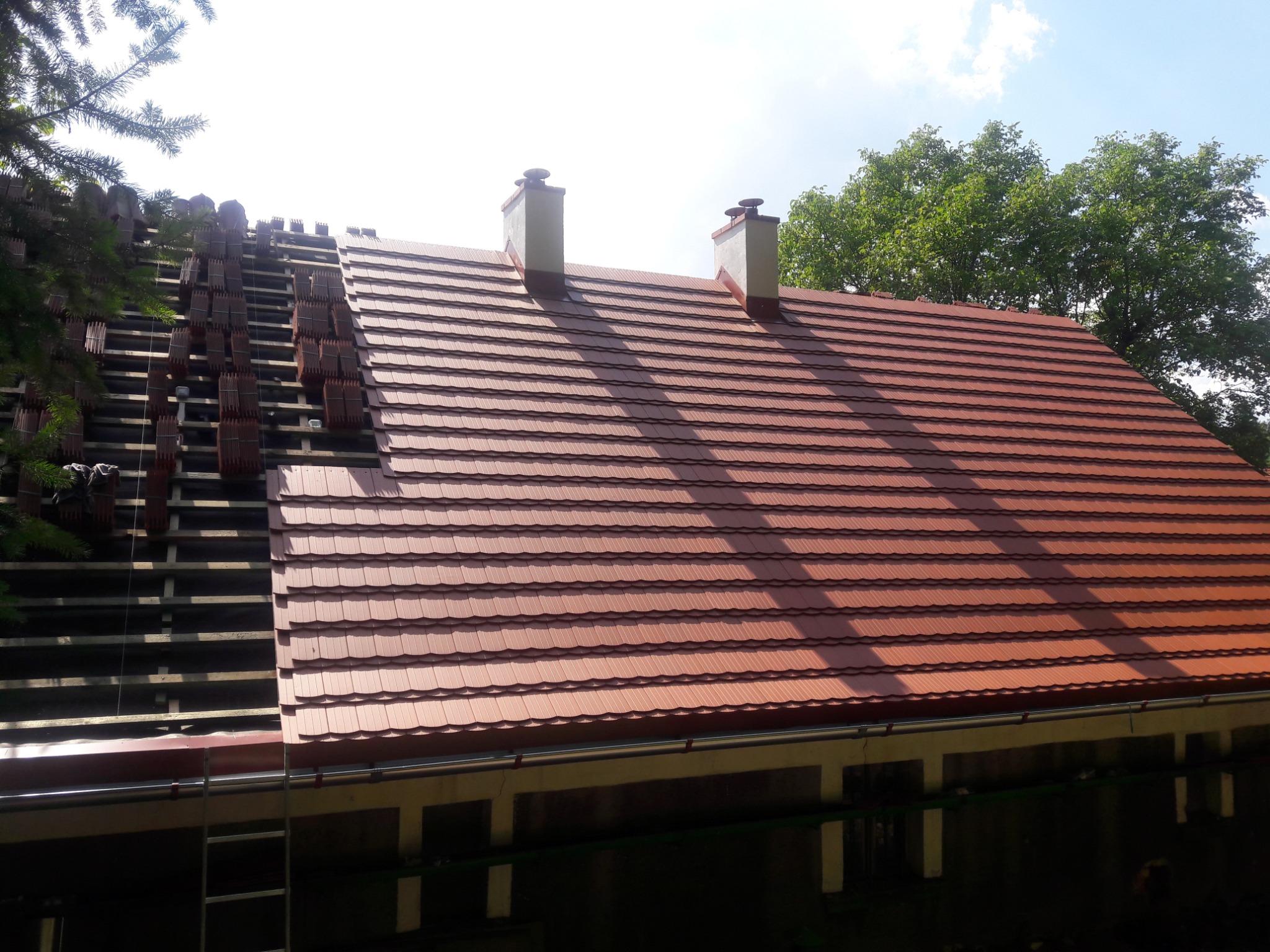 Częściowo pokryty dach z czerwonej dachówki ceramicznej, widoczna więźba dachowa i kominy, prace dekarskie w toku.