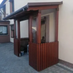 Drewniana weranda z przeszklonymi ścianami i niskim płotkiem, w kolorze ciemnego brązu, przylegająca do beżowego budynku z szarym brukowanym chodnikiem.