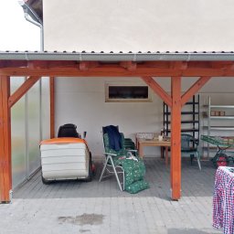 Stolarstwo - Zadaszenie drewniane z pomalowanymi na brązowo elementami konstrukcyjnymi, pod którym stoi kosiarka, krzesła ogrodowe, stół z naczyniami i wózek z narzędziami.