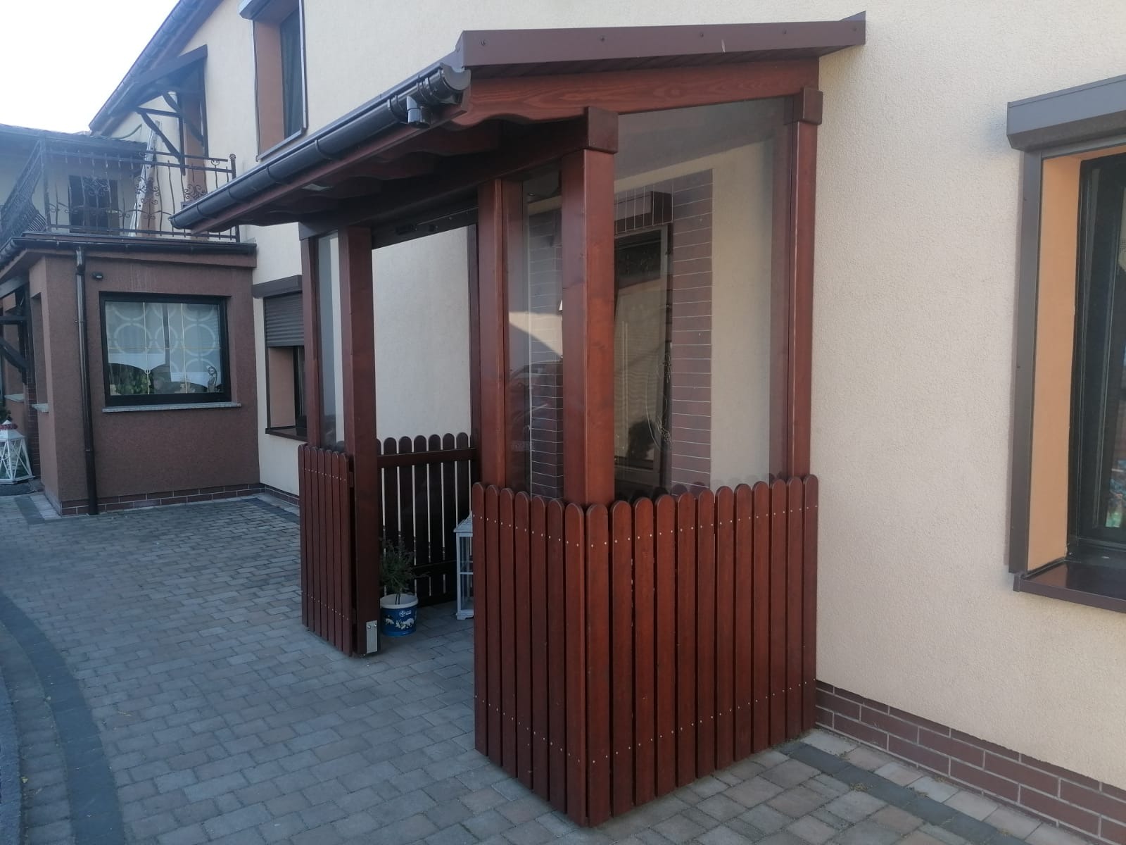 Drewniana weranda z przeszklonymi ścianami i niskim płotkiem, w kolorze ciemnego brązu, przylegająca do beżowego budynku z szarym brukowanym chodnikiem.