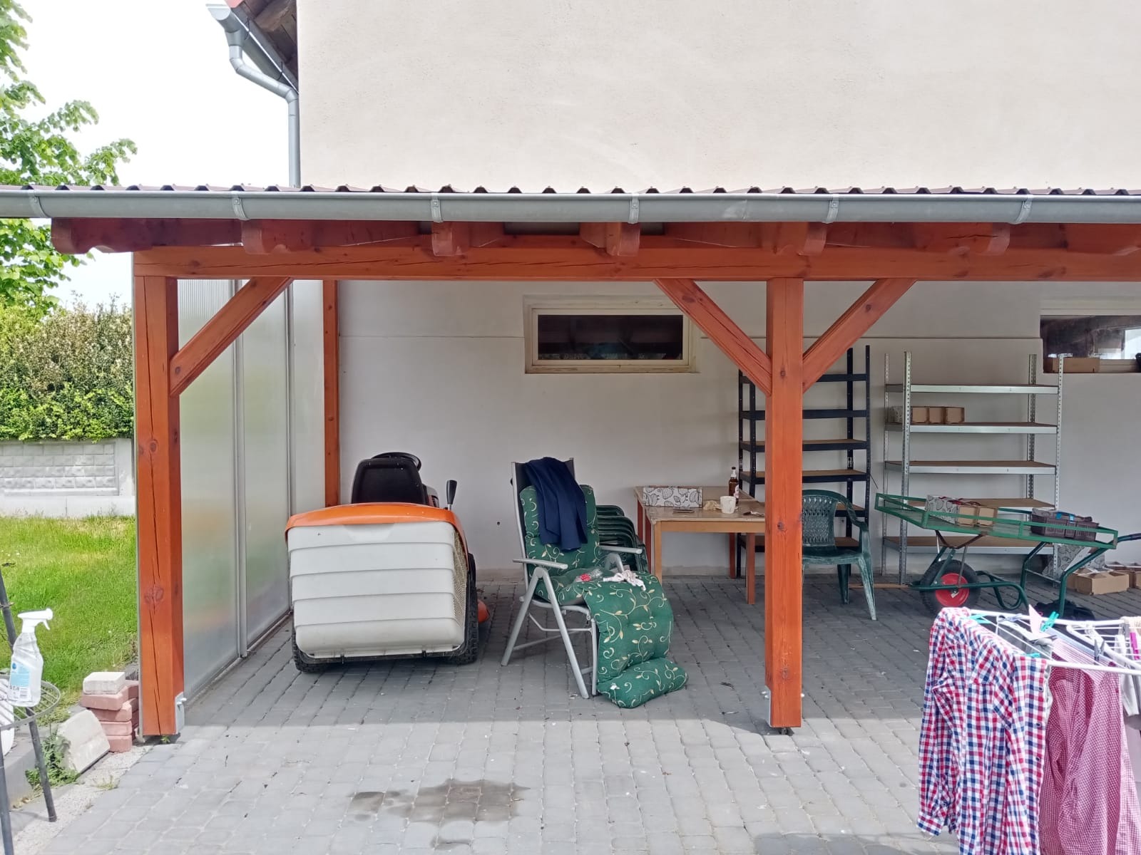 Zadaszenie drewniane z pomalowanymi na brązowo elementami konstrukcyjnymi, pod którym stoi kosiarka, krzesła ogrodowe, stół z naczyniami i wózek z narzędziami.