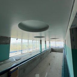 RAJ MARCIN ŚWIDERSKI - Wnętrze w trakcie prac wykończeniowych: sufit z okrągłymi wnękami, ściany częściowo obłożone płytami, widoczne okna i elementy konstrukcyjne.