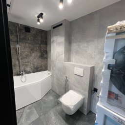 RAJ MARCIN ŚWIDERSKI - Nowoczesna łazienka po remoncie: szare płytki imitujące marmur na ścianach i podłodze, narożna biała wanna, podwieszana toaleta z przyciskiem, oświetlenie punktowe, pralka w folii ochronnej po prawej.