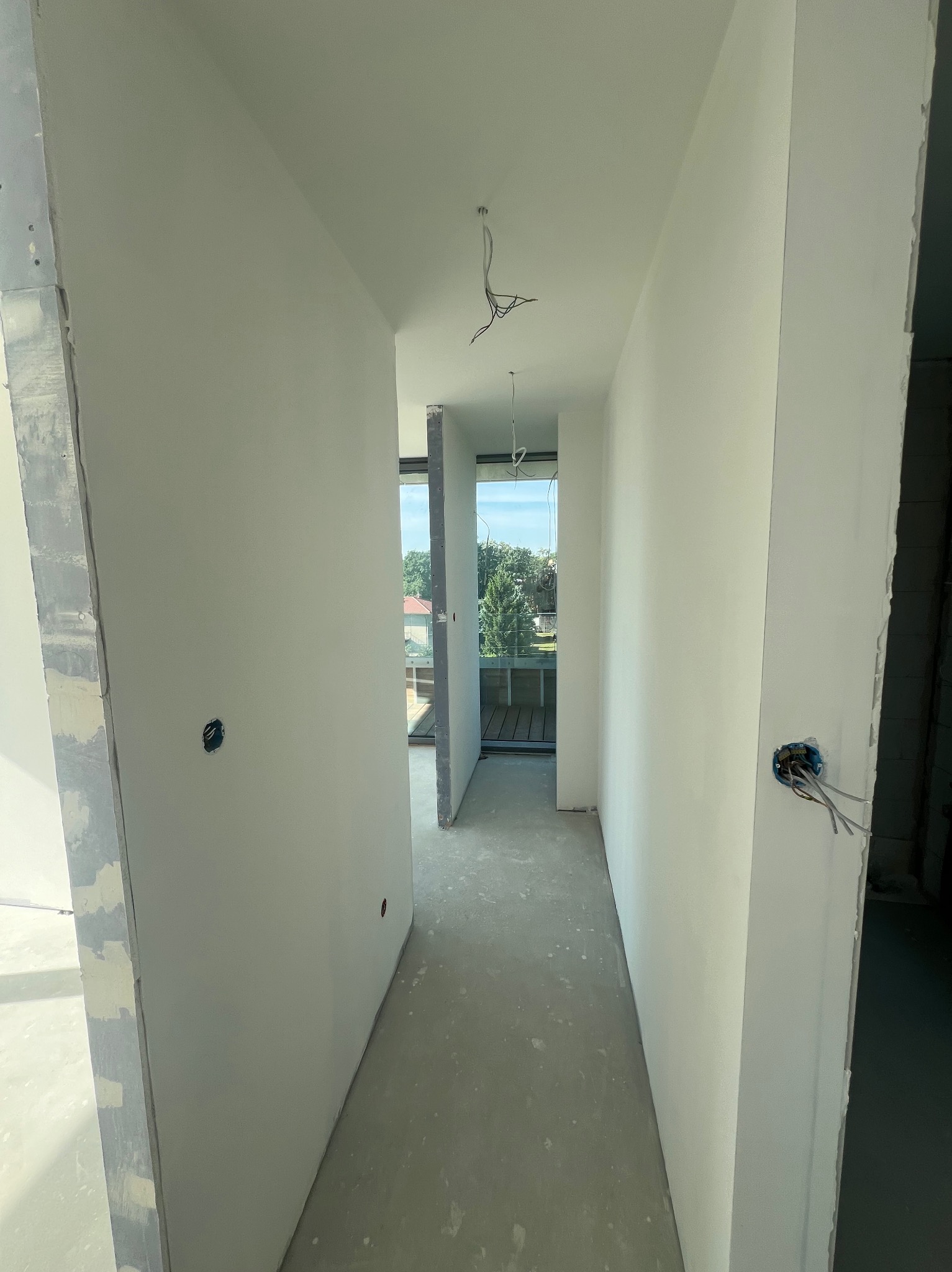 Korytarz w trakcie prac wykończeniowych, ściany po szpachlowaniu i malowaniu na biało, widoczne puszki elektryczne z wystającymi przewodami, surowy beton na podłodze, w tle widok na zieleń przez...