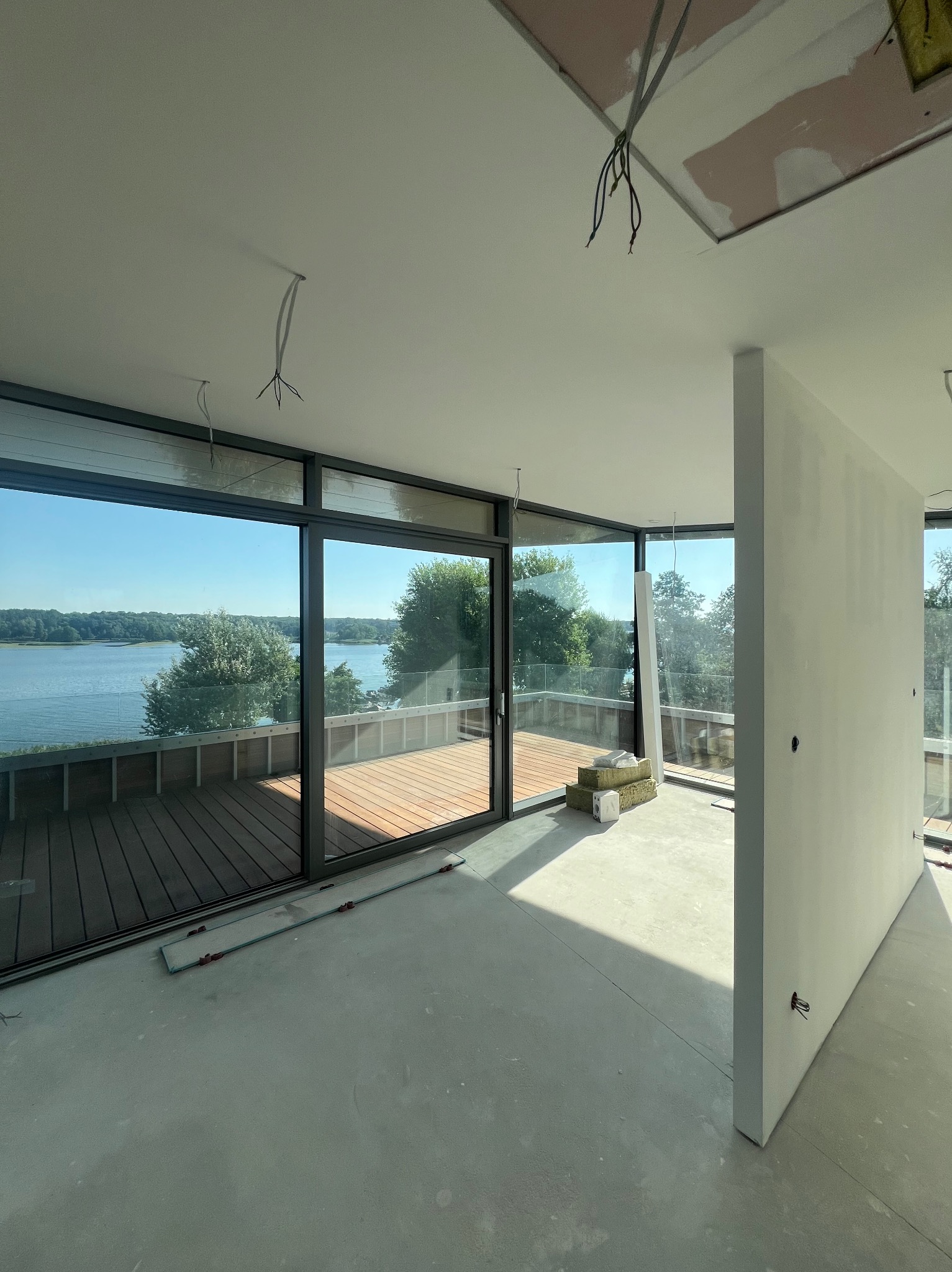 Wnętrze w trakcie prac wykończeniowych, widoczne duże okna z widokiem na jezioro, ściany przygotowane do malowania, wystające kable elektryczne z sufitu, materiały izolacyjne na podłodze.