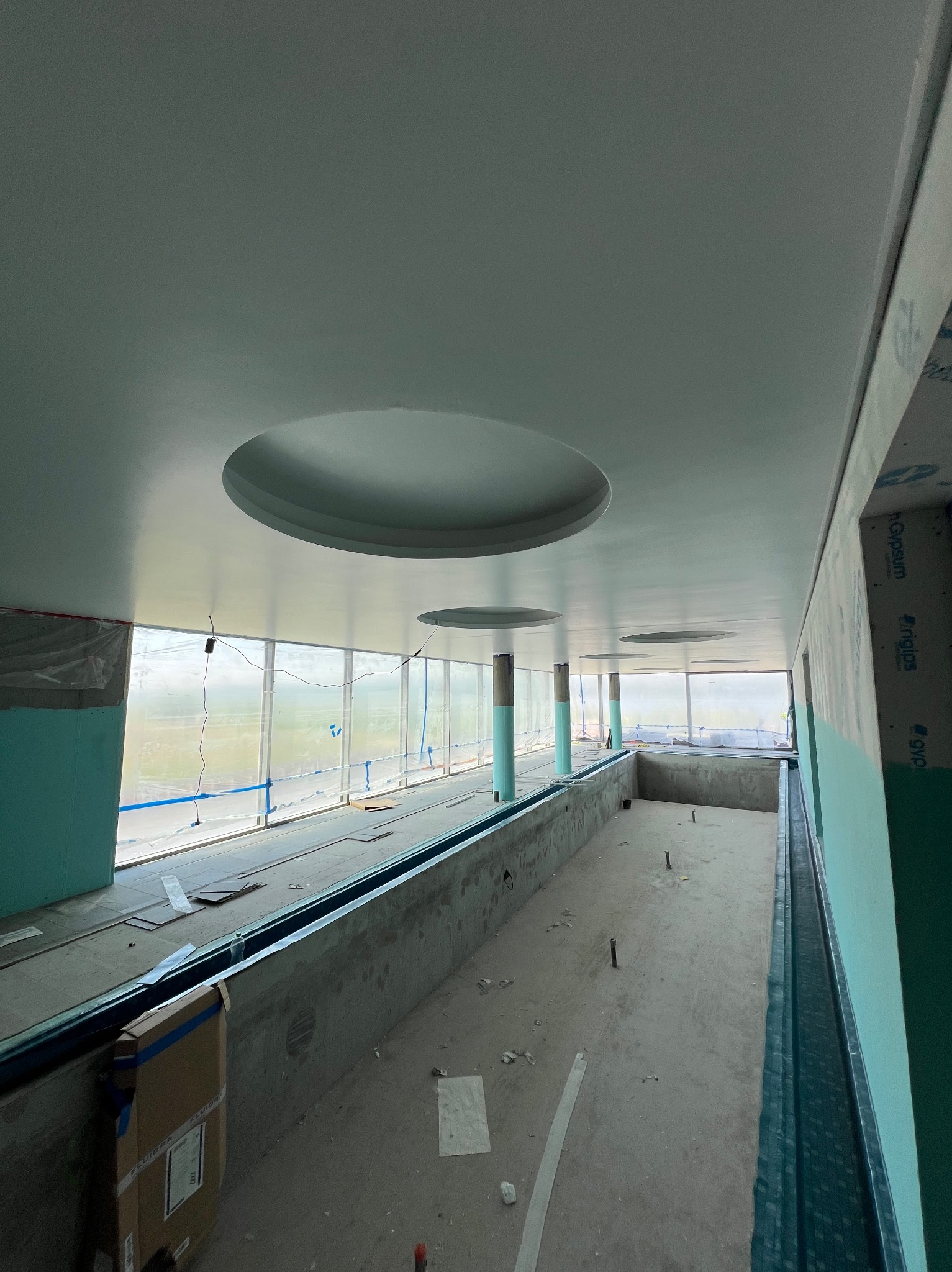 Wnętrze w trakcie prac wykończeniowych: sufit z okrągłymi wnękami, ściany częściowo obłożone płytami, widoczne okna i elementy konstrukcyjne.