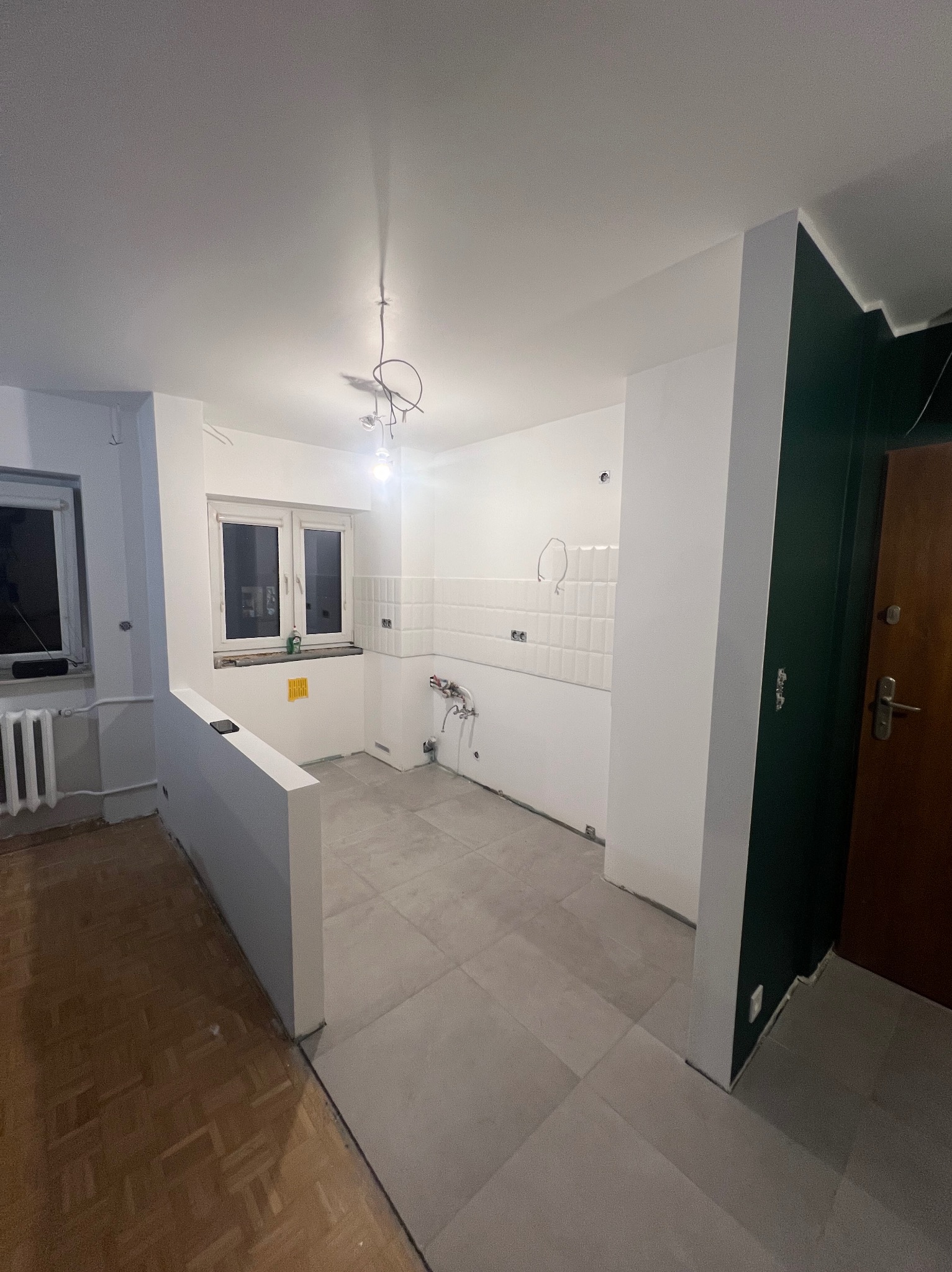 Wnętrze w trakcie prac: białe ściany, szare płytki podłogowe, częściowo położone białe płytki na ścianie w kuchni, wystające kable, widoczny fragment parkietu w pokoju obok.