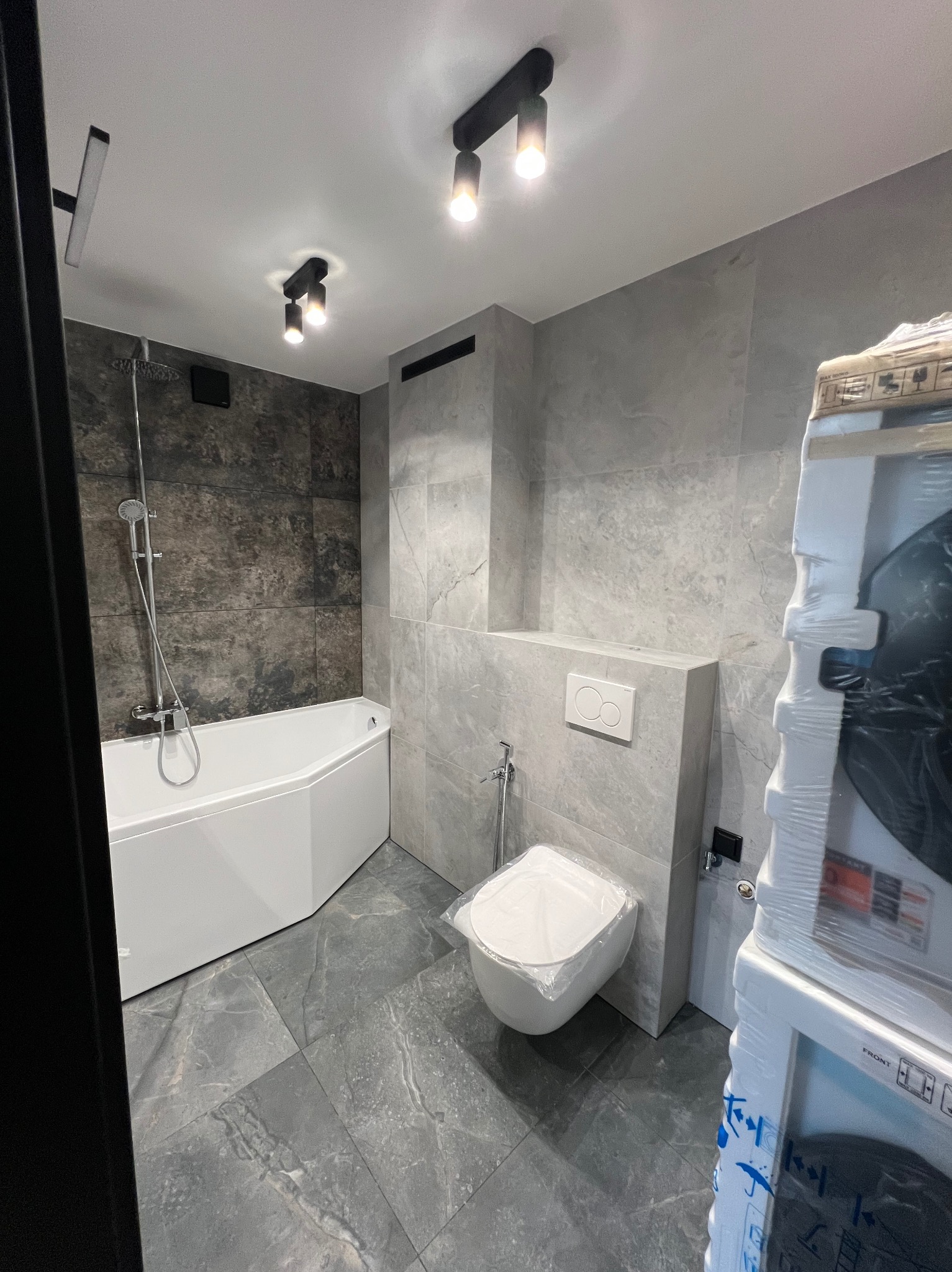 Nowoczesna łazienka po remoncie: szare płytki imitujące marmur na ścianach i podłodze, narożna biała wanna, podwieszana toaleta z przyciskiem, oświetlenie punktowe, pralka w folii ochronnej po prawej.