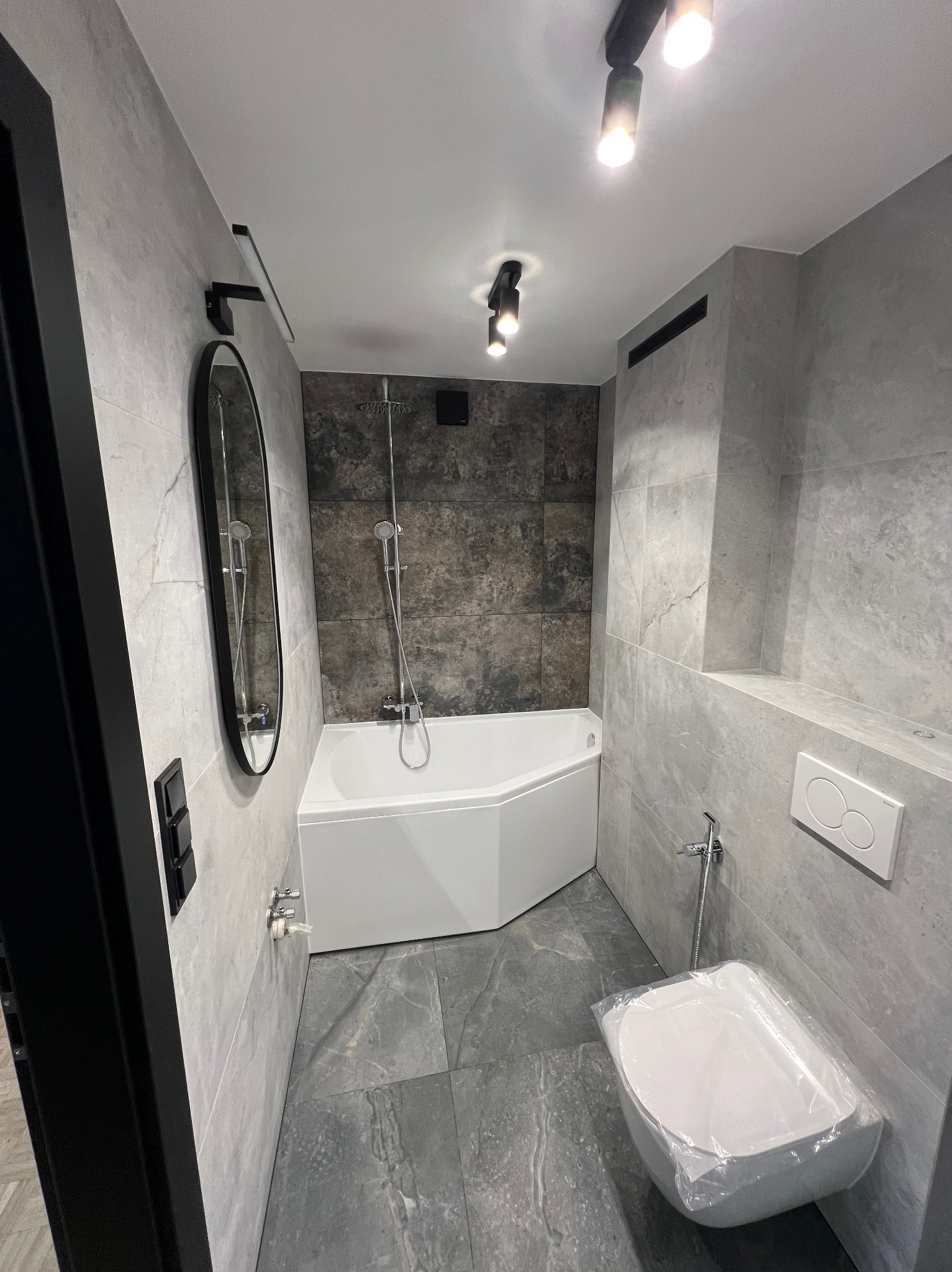 Nowoczesna łazienka w szarościach z narożną wanną, owalnym lustrem i podwieszaną toaletą w folii ochronnej, oświetlona punktowymi lampami.