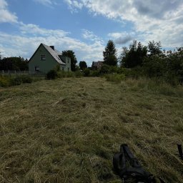 Krystian Jakubowski - Świeżo skoszone pole trawy z widocznymi śladami cięcia, w tle zielony dom i błękitne niebo z chmurami. Częściowo widoczny sprzęt do koszenia w prawym dolnym rogu.