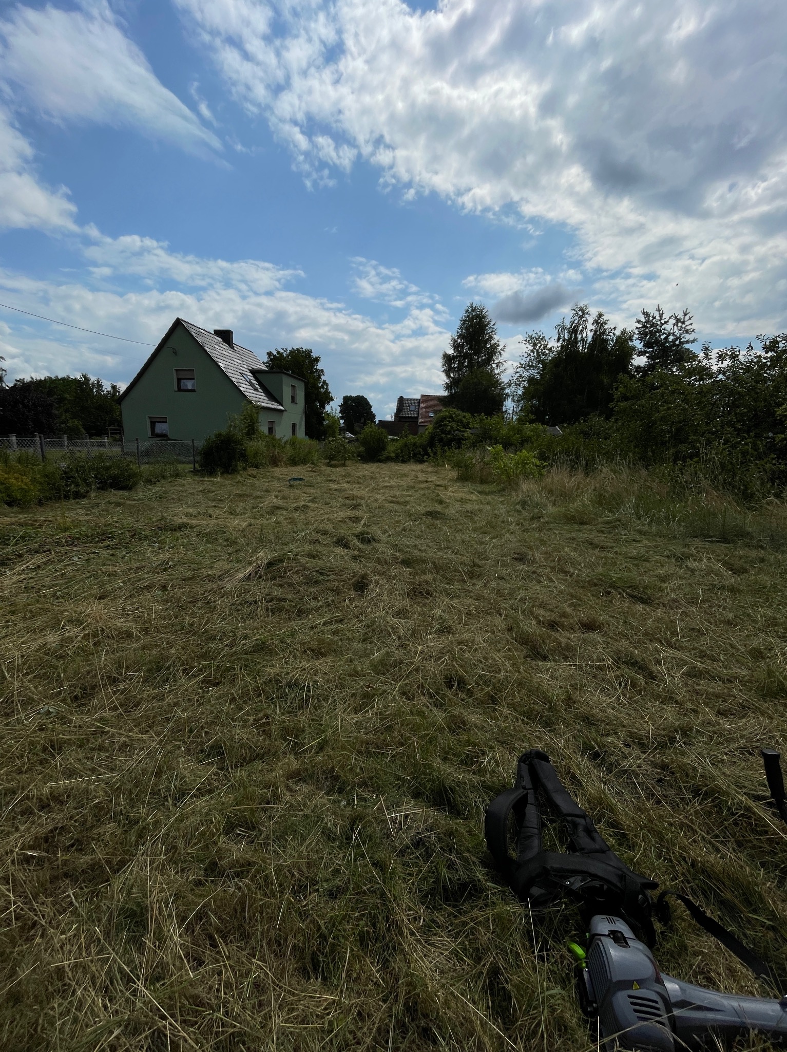 Świeżo skoszone pole trawy z widocznymi śladami cięcia, w tle zielony dom i błękitne niebo z chmurami. Częściowo widoczny sprzęt do koszenia w prawym dolnym rogu.