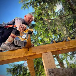 Republika Wolności Sp. z o.o. - Monter w trakcie budowy drewnianej konstrukcji, prawdopodobnie tarasu lub wiaty, w tle widoczne drzewa i niebo, używa wkrętarki Dewalt.