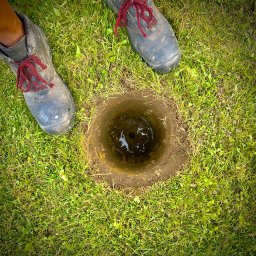 Republika Wolności Sp. z o.o. - Wykopany otwór w trawie, prawdopodobnie pod słupek, obok stoją buty robocze z czerwonymi sznurówkami.