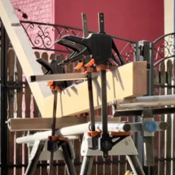 Drewniana konstrukcja w trakcie montażu, zabezpieczona czarnymi ściskami stolarskimi, ustawiona na białych kozłach montażowych na tle czerwonej ściany i metalowego ogrodzenia.