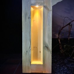 Republika Wolności Sp. z o.o. - Drewniana kolumna oświetleniowa z wbudowanym panelem LED, widoczna struktura drewna i ciepłe światło na tle ciemnego otoczenia.