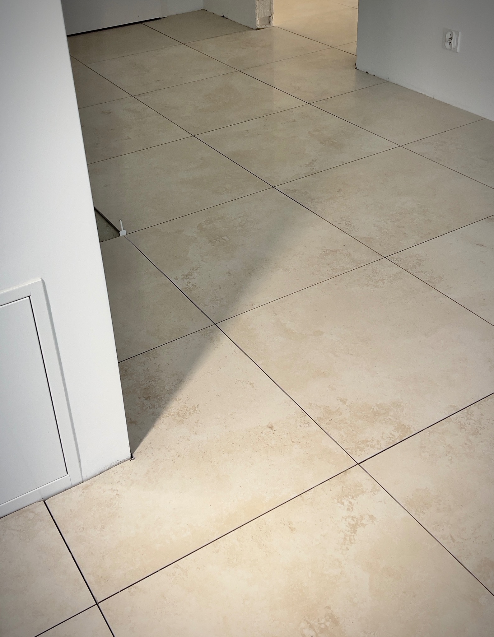 Beżowe płytki podłogowe w nowym apartamencie, widoczny fragment ściany z gniazdkiem elektrycznym i biała listwa przypodłogowa.