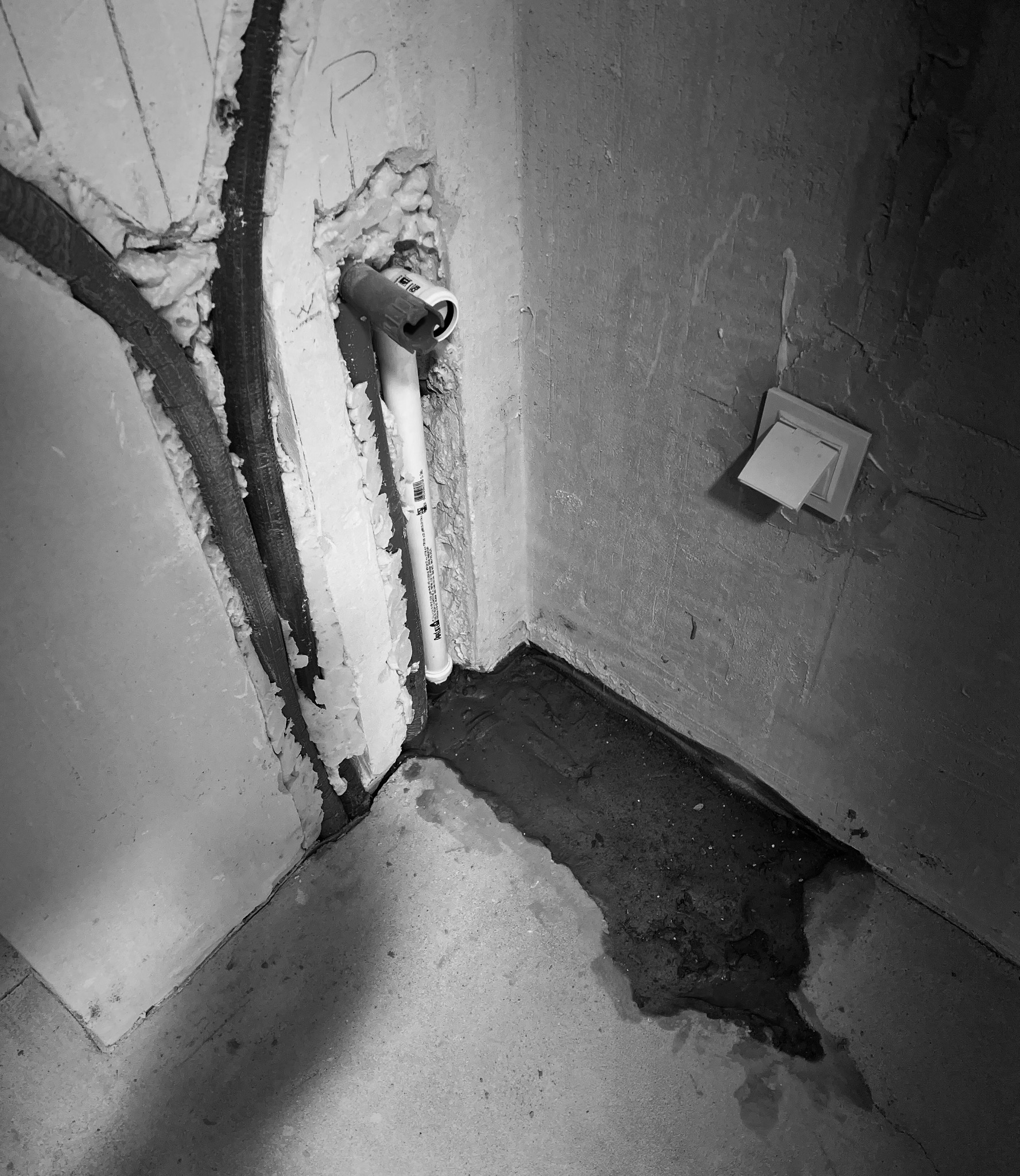 Czarno-białe zdjęcie przedstawia fragment ściany w stanie surowym z wystającą rurą PCV uszczelnioną pianką montażową, widoczne ślady wilgoci na podłodze i wystający kabel elektryczny.