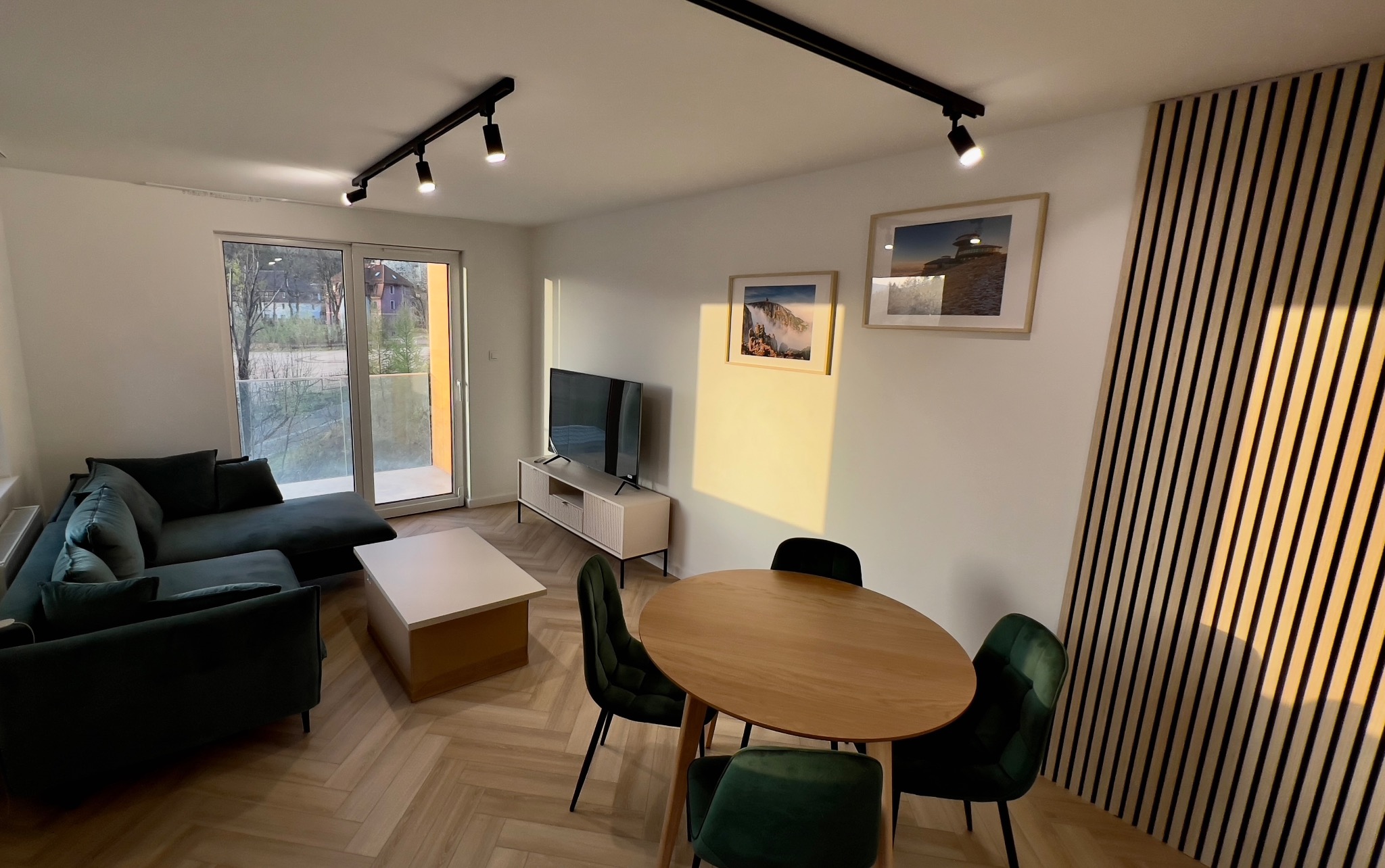 Nowoczesny salon w stanie deweloperskim z zieloną sofą narożną, drewnianym stołem jadalnym i panelami dekoracyjnymi na ścianie, oświetlony reflektorami sufitowymi.