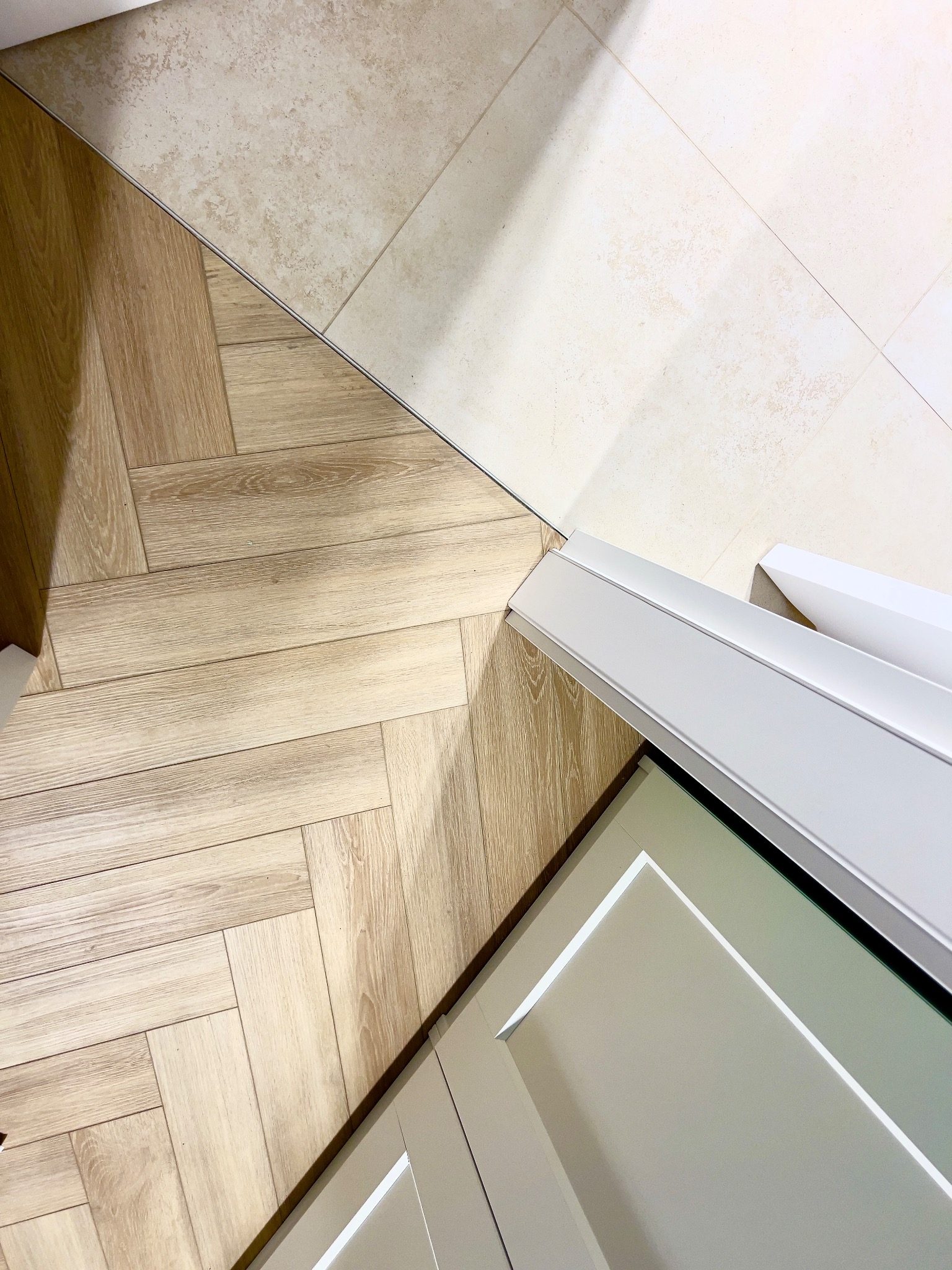 Połączenie podłogi drewnopodobnej ułożonej w jodełkę z jasnymi płytkami ceramicznymi, widoczna dolna część białych drzwi.