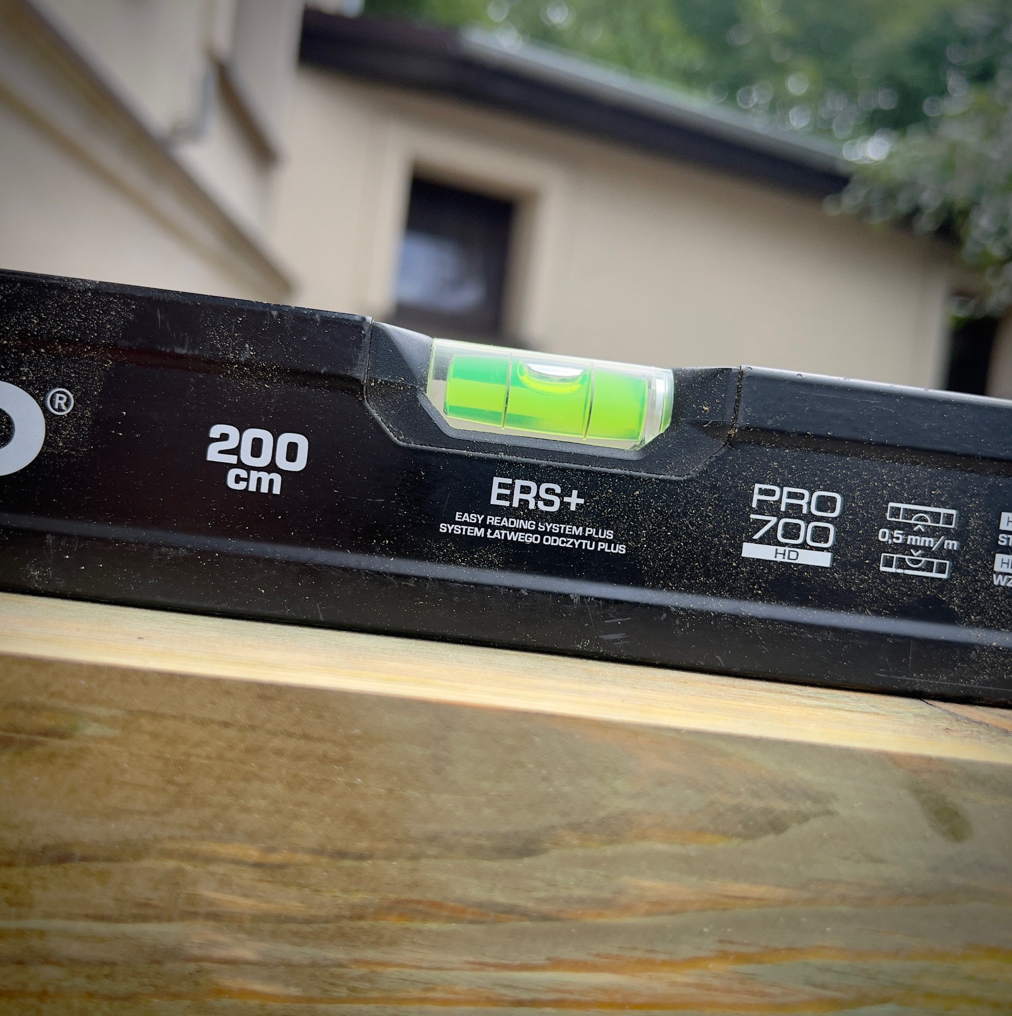 Poziomnica o długości 200 cm, model PRO 700 HD, leżąca na surowym, jasnym drewnie na tle budynku z oknem i zielenią.
