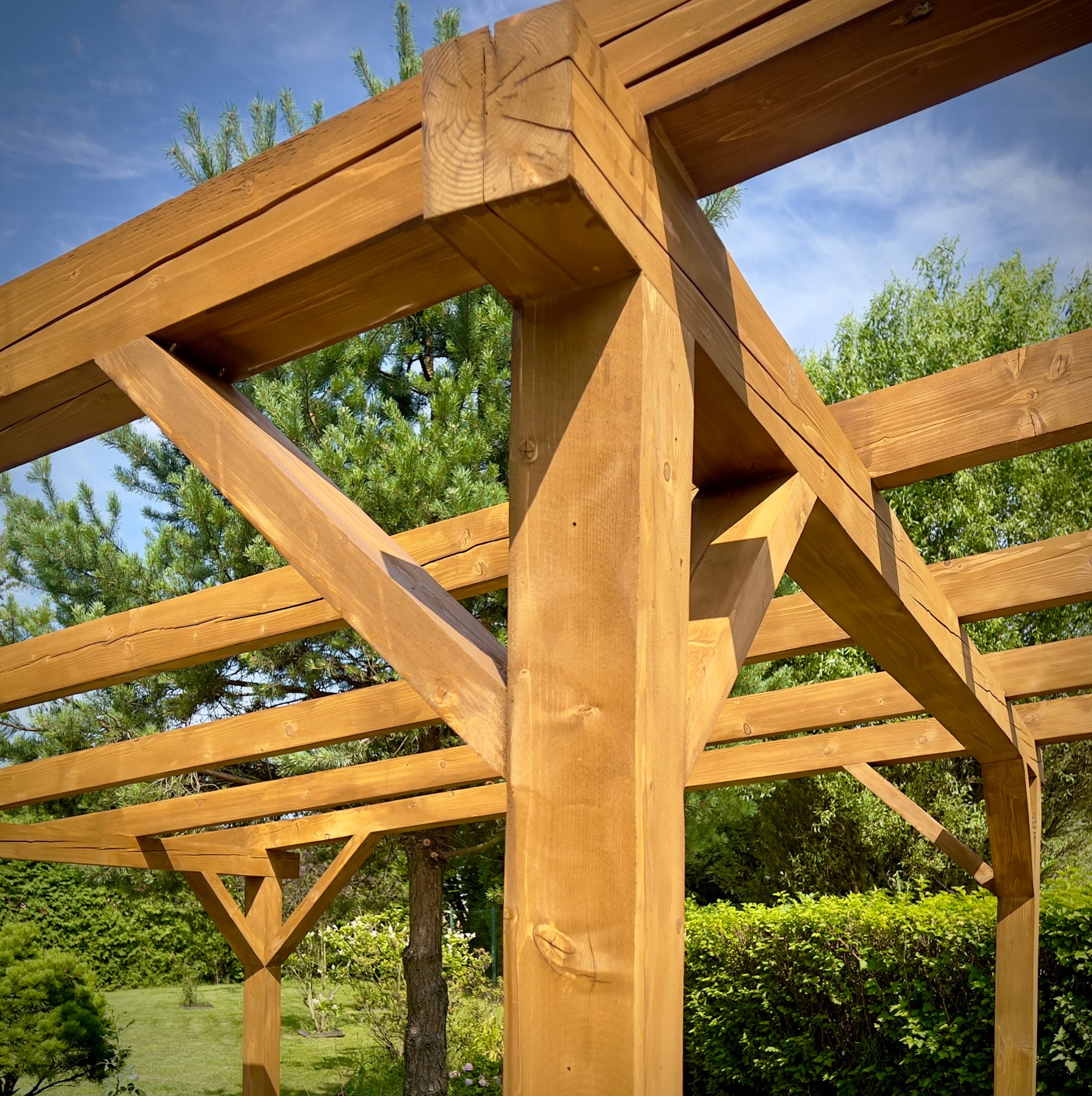 Drewniana konstrukcja wiaty ogrodowej z belek o przekroju kwadratowym, widoczne połączenia ciesielskie i naturalne otoczenie zieleni.