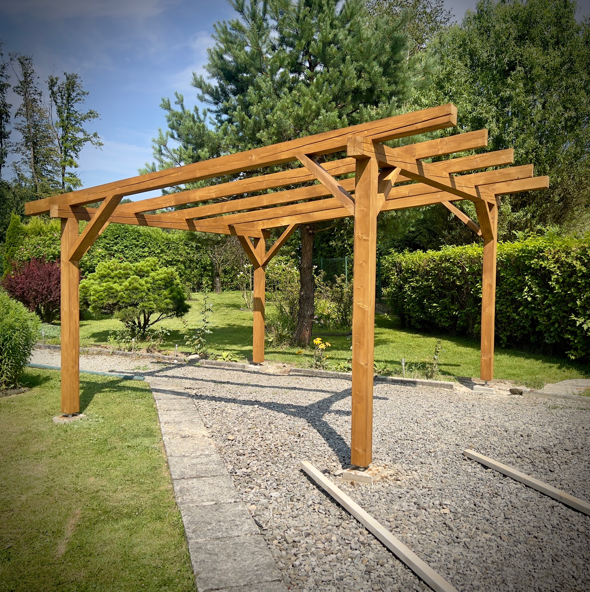 Drewniana konstrukcja wiaty z belek na fundamencie słupowym, widoczne kotwy z regulacją wysokości, tło z zieleni ogrodowej i błękitnego nieba.