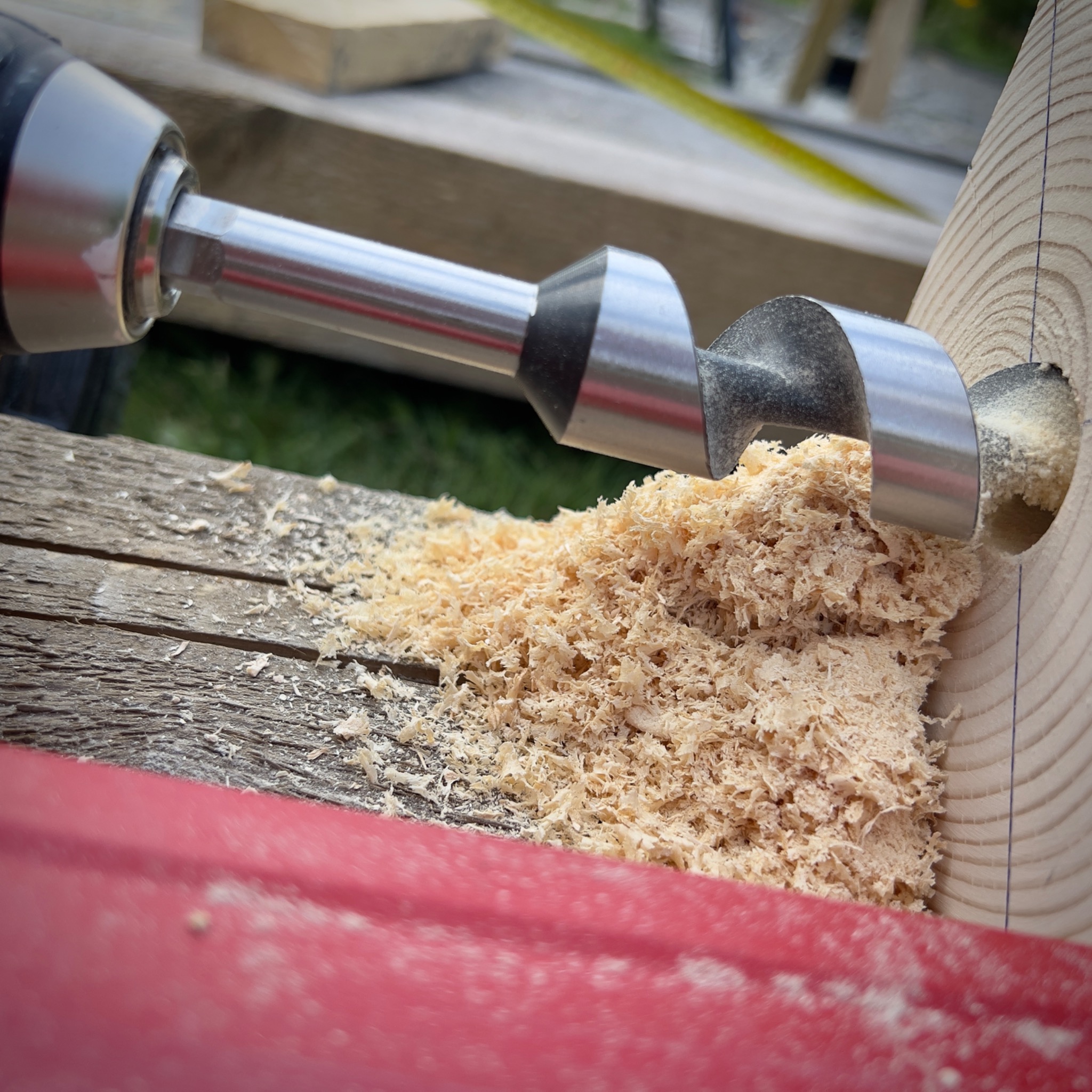 Wiertło świdrowe w trakcie wiercenia otworu w drewnianym słupku, z widocznymi wiórami i linią pomocniczą wyznaczoną na drewnie.