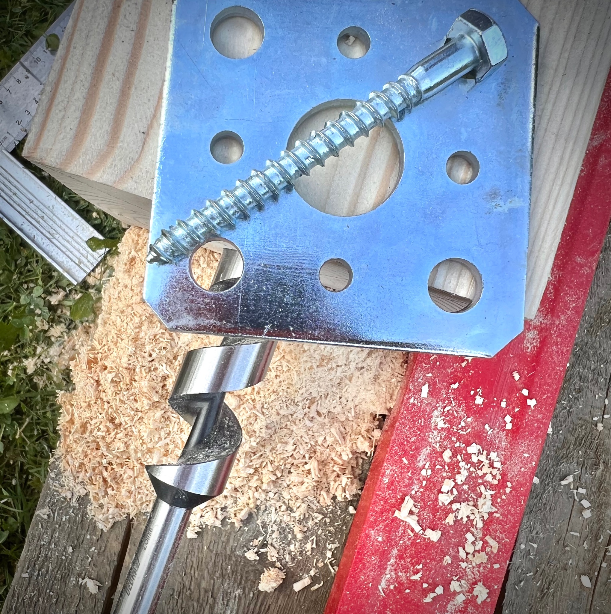 Metalowa płytka mocująca z otworami, wkręt do drewna, wiertło świdrowe, fragment deski, miarka i wióry na tle trawy.