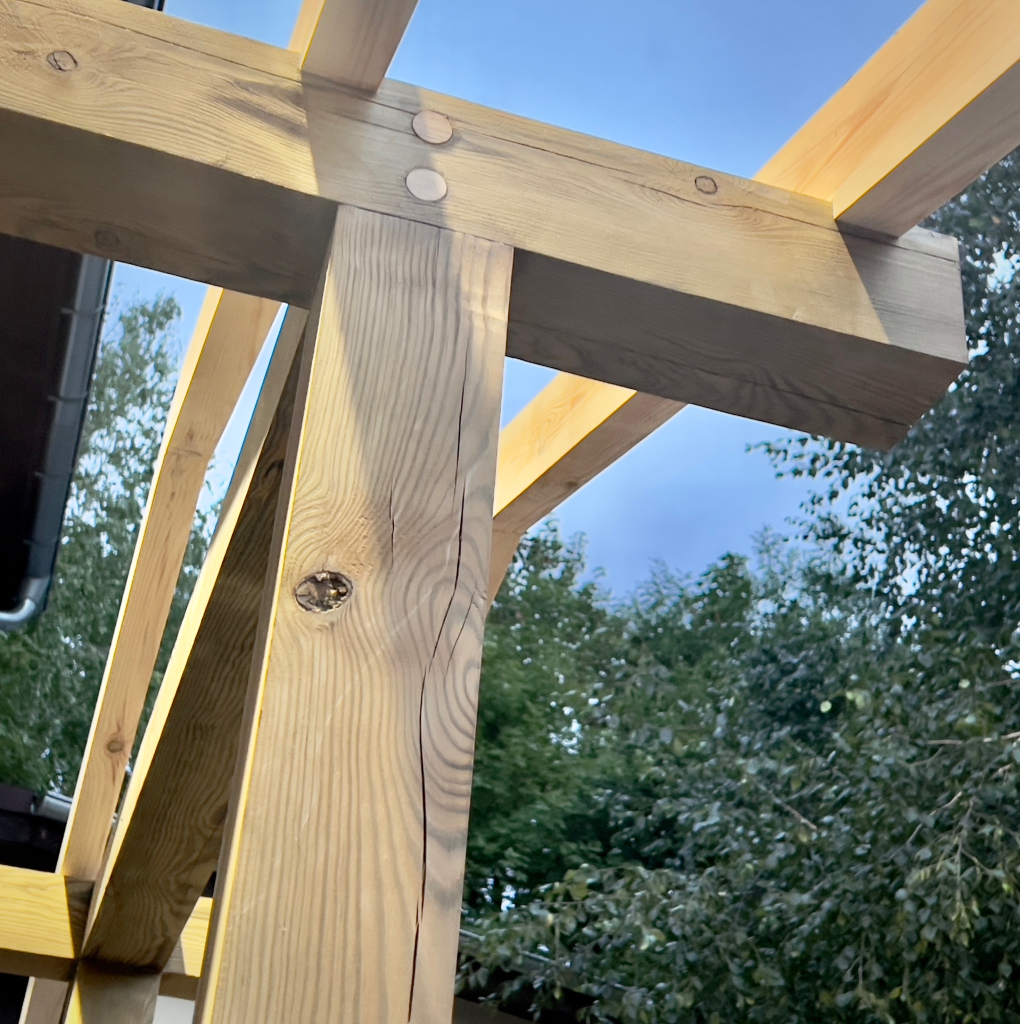 Fragment drewnianej konstrukcji tarasu w trakcie budowy, widoczne połączenia belek z użyciem drewnianych kołków, tło z zieleni i fragment budynku.