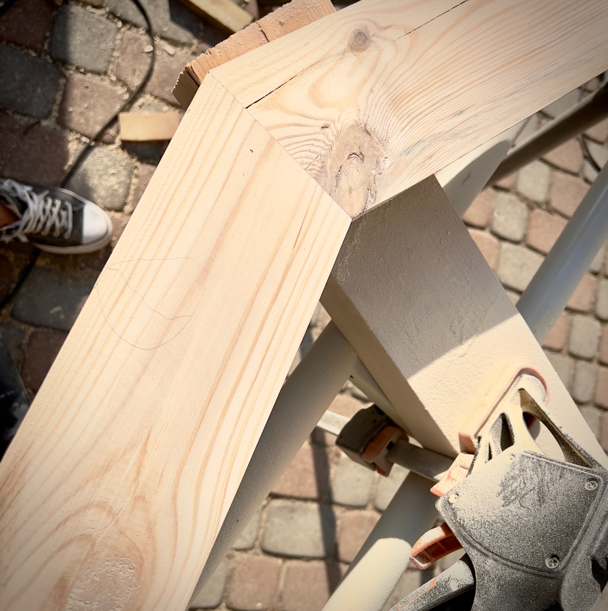 Połączenie dwóch belek z jasnego drewna, zabezpieczone ściskiem stolarskim, na tle kostki brukowej i fragmentu buta.