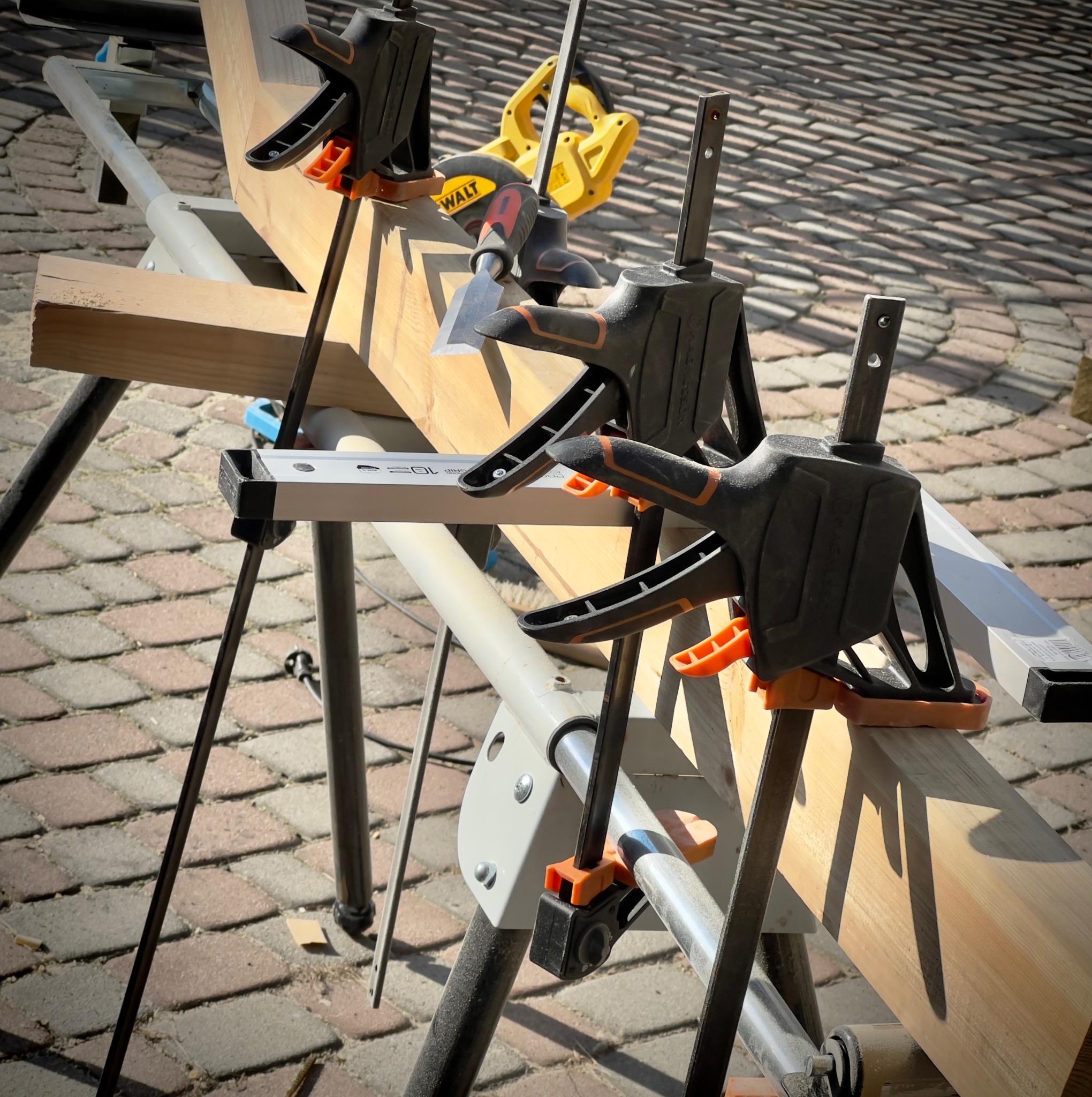 Deska mocowana do stojaka za pomocą ścisków stolarskich; w tle widoczna piła tarczowa i dłuto. Praca w toku, na zewnątrz, na brukowanym podłożu.