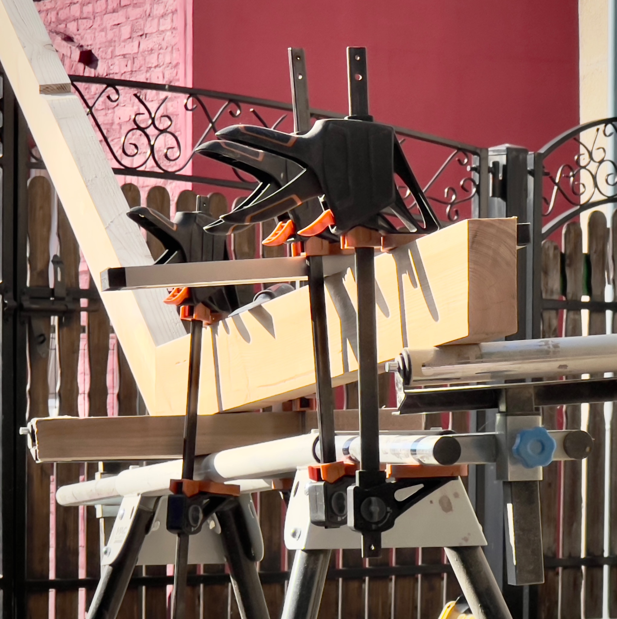 Drewniana konstrukcja w trakcie montażu, zabezpieczona czarnymi ściskami stolarskimi, ustawiona na białych kozłach montażowych na tle czerwonej ściany i metalowego ogrodzenia.
