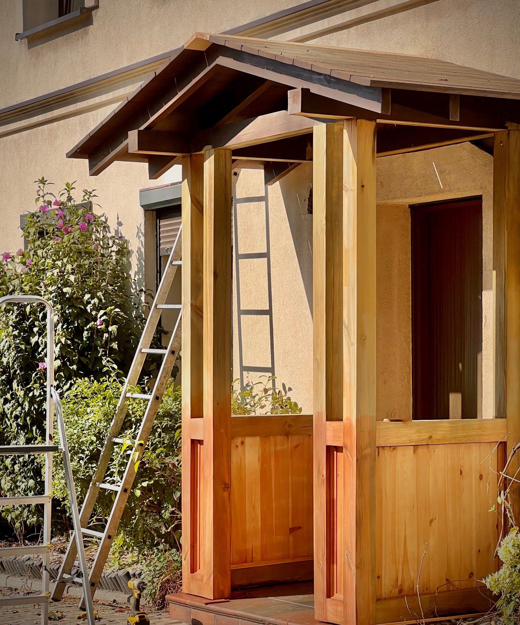 Wznoszona drewniana konstrukcja ganku z widocznymi elementami szkieletu, dwiema drabinami opartymi o ścianę budynku i wkrętarką leżącą na ziemi, sugerująca trwające prace wykończeniowe.