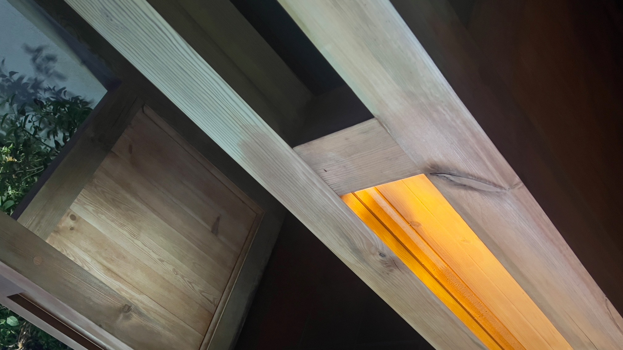 Drewniana konstrukcja ganku w trakcie renowacji, widoczne elementy oświetlenia i fragment roślinności w tle.