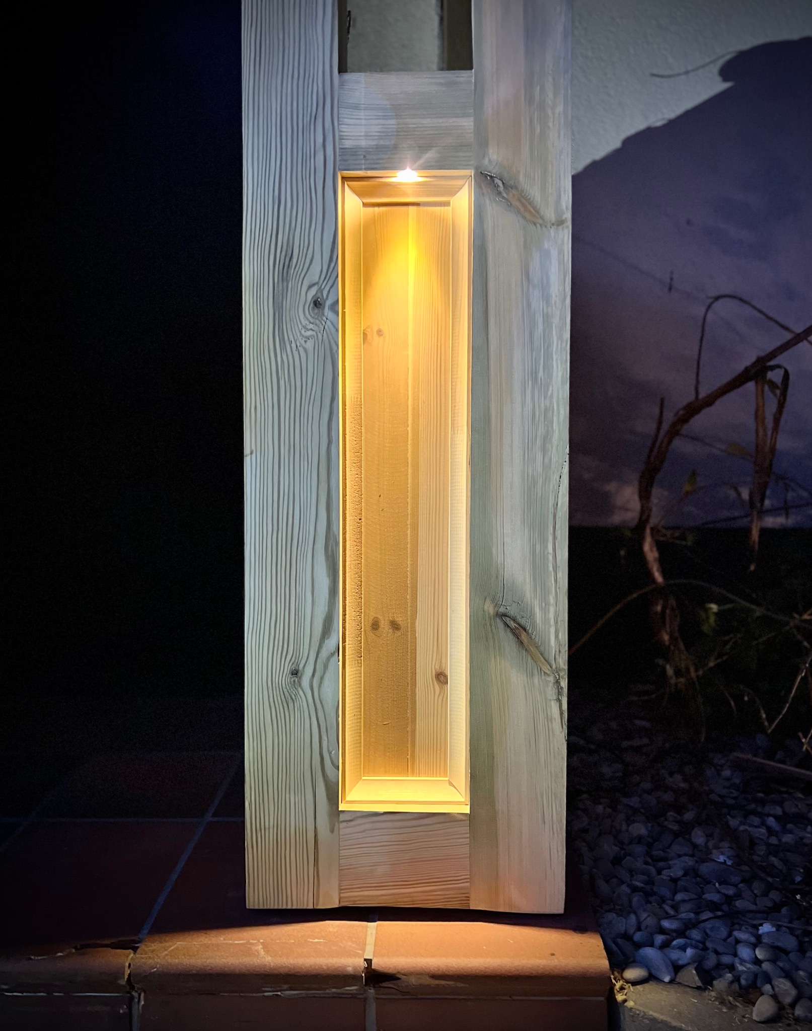 Drewniana kolumna oświetleniowa z wbudowanym panelem LED, widoczna struktura drewna i ciepłe światło na tle ciemnego otoczenia.