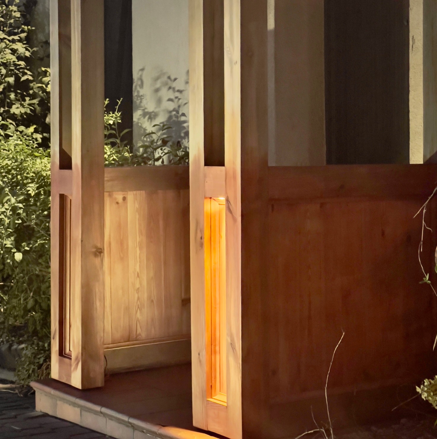 Drewniana konstrukcja ganku oświetlona od wewnątrz, widoczne fragmenty roślinności w tle, nocna sceneria.
