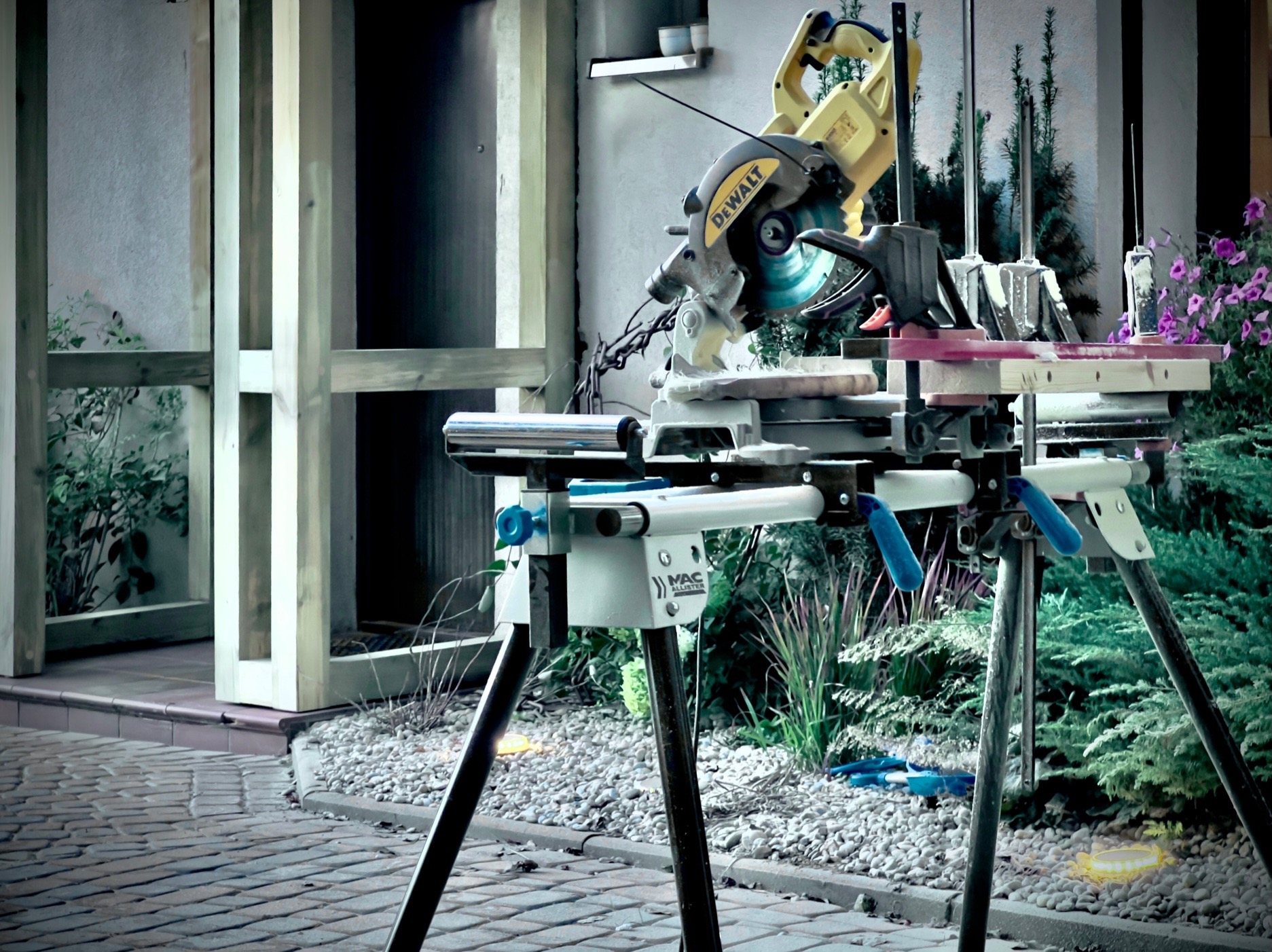 Ustawiona na zewnątrz piła ukośnica DeWalt na stojaku Mac Allister, przygotowana do cięcia drewna, w tle fragment drewnianej konstrukcji i roślinność ogrodowa.