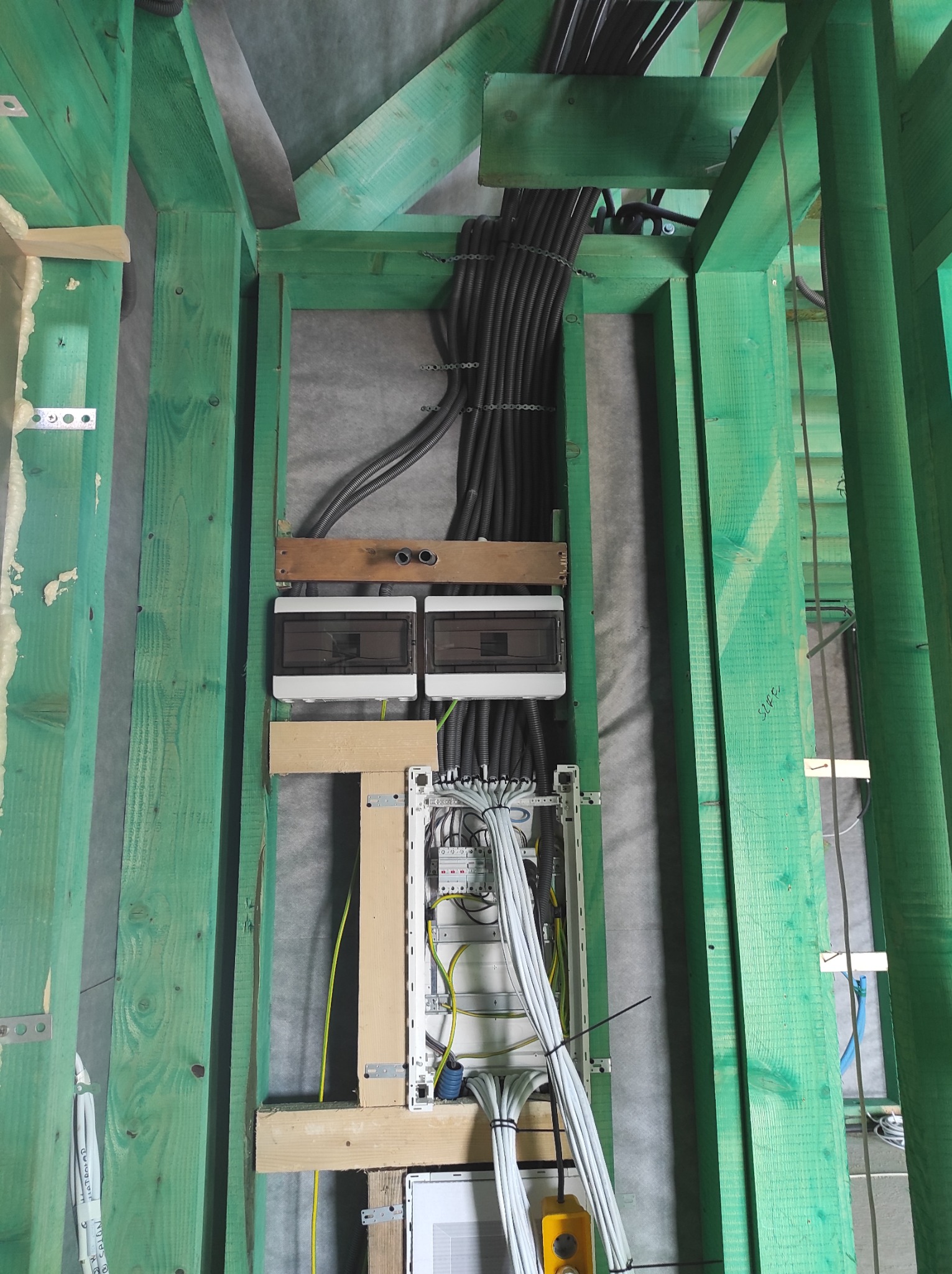 Rozdzielnica elektryczna z licznymi przewodami wewnątrz konstrukcji drewnianej pomalowanej na zielono, widoczne także peszle z kablami, całość w surowym stanie wykończenia.