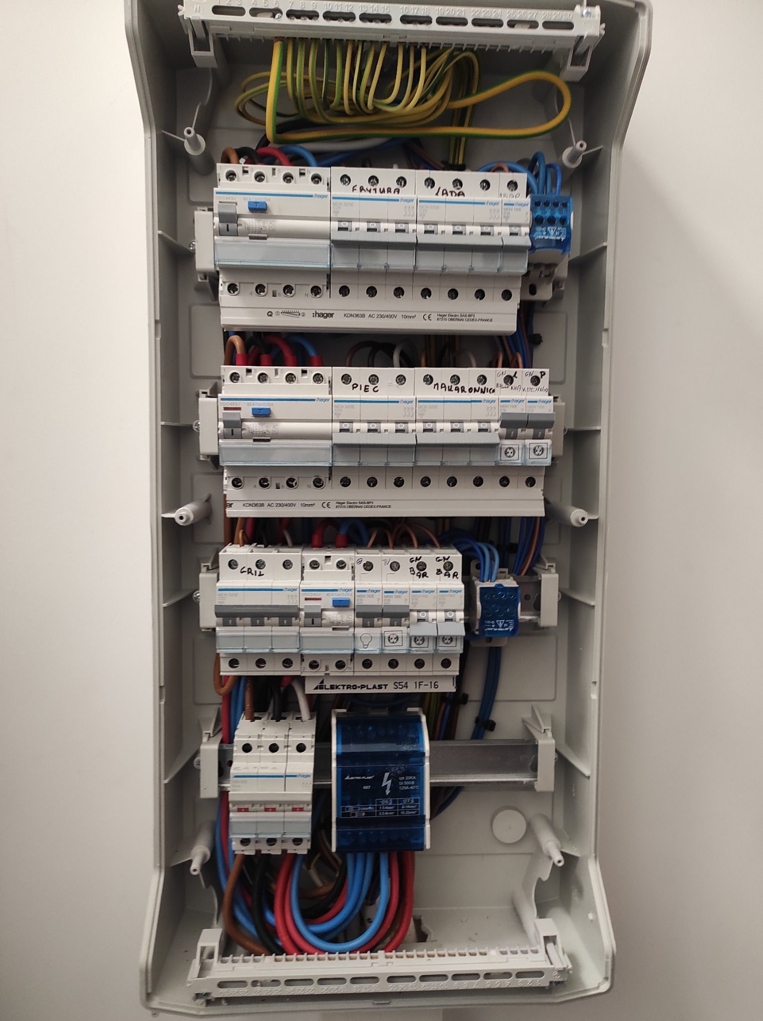 Otwarta, szara skrzynka rozdzielcza z widocznymi bezpiecznikami, przewodami w kolorach: żółto-zielonym, niebieskim, czerwonym i brązowym; z odręcznymi napisami na obudowach bezpieczników: 'Frytura'...