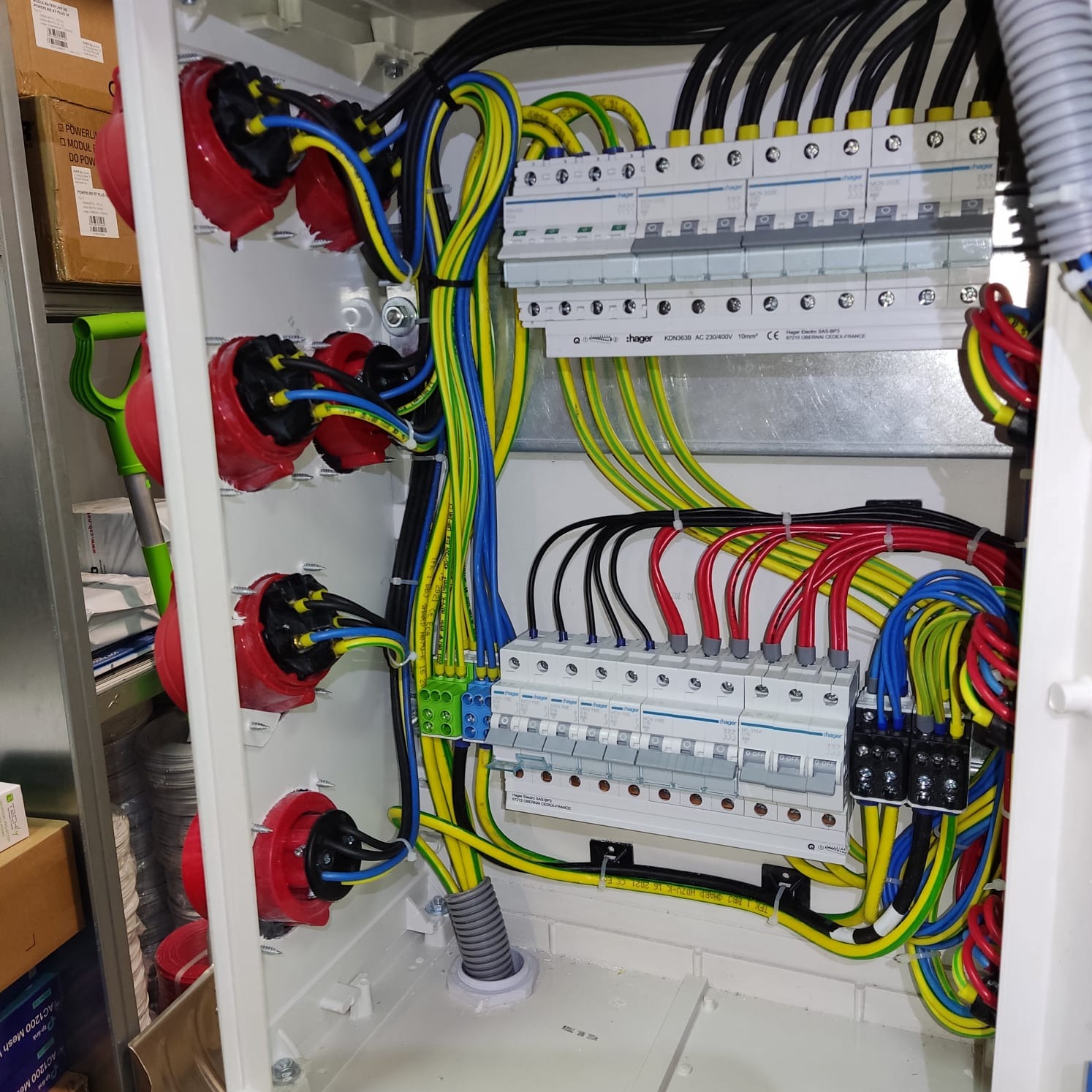 Wnętrze rozdzielnicy elektrycznej z widocznymi bezpiecznikami, przewodami w kolorach niebieskim, żółto-zielonym, czarnym i czerwonym, oraz gniazdami siłowymi z czerwonymi obudowami. Widoczny...