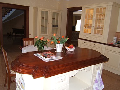 Klasyczna kuchnia z wyspą o ciemnym, drewnianym blacie, na której leżą gazety, a w tle widoczne przeszklone szafki i kwiaty w wazonie.