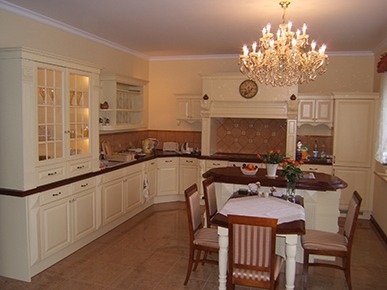 Klasyczna kuchnia w odcieniach beżu z wyspą, drewnianym blatem i kryształowym żyrandolem nad stołem z czterema krzesłami.