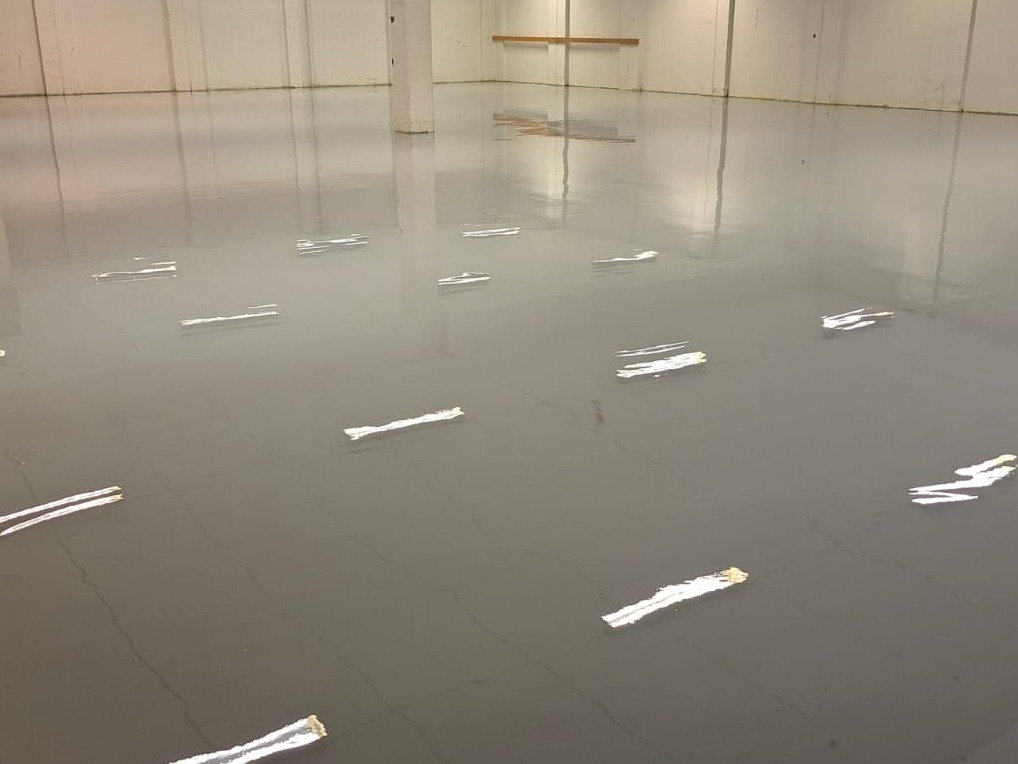 Gładka, szara powierzchnia w hali z refleksami światła, prawdopodobnie świeżo wylana. Widoczne pęknięcia i niedoskonałości w strukturze.